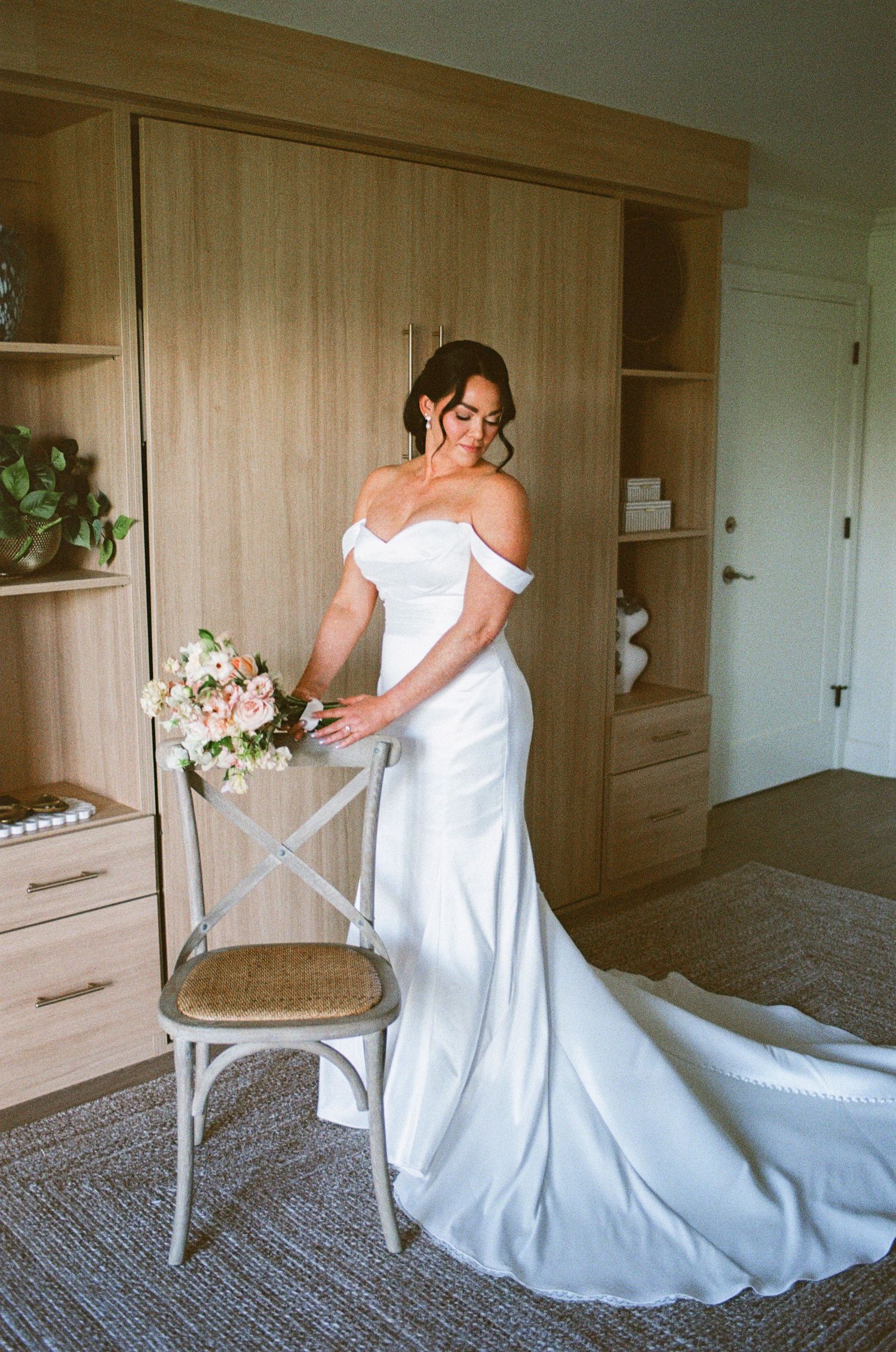 Bride in off-shoulder white gown with train holds bouquet beside wooden chair in elegant room.