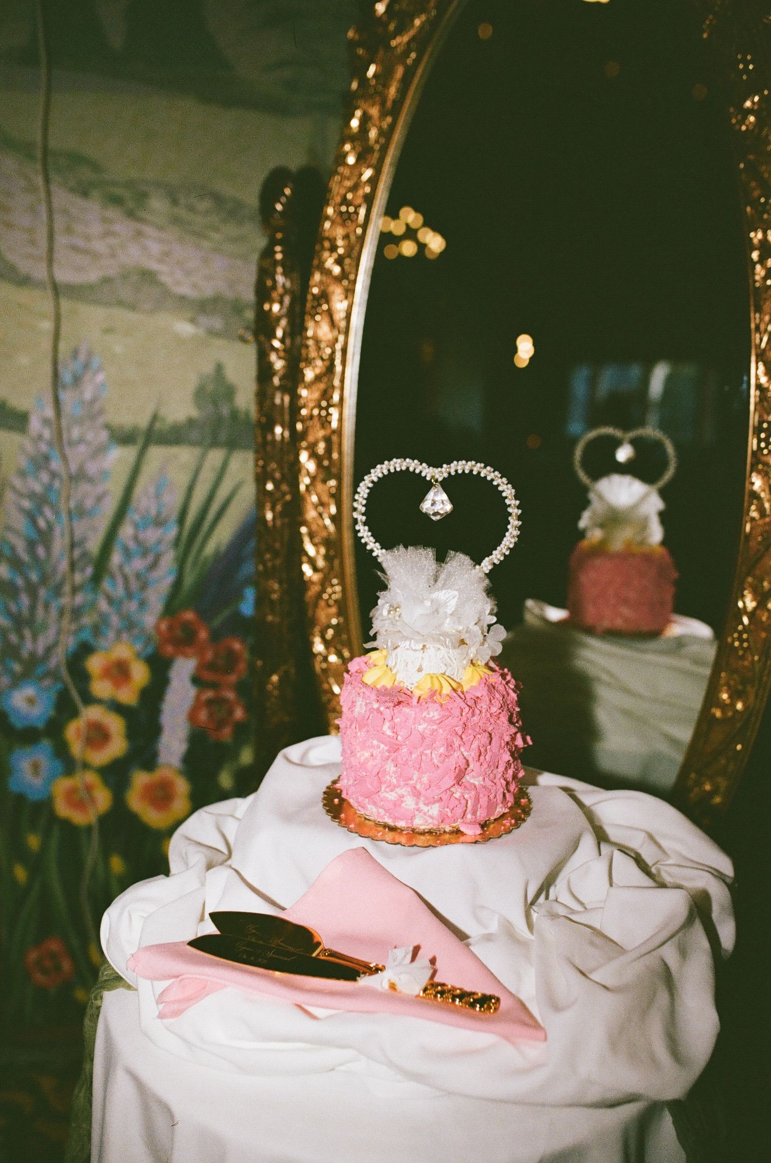 Pink rosette wedding cake with crystal heart topper on white table, reflected in ornate gold mirror with floral backdrop.