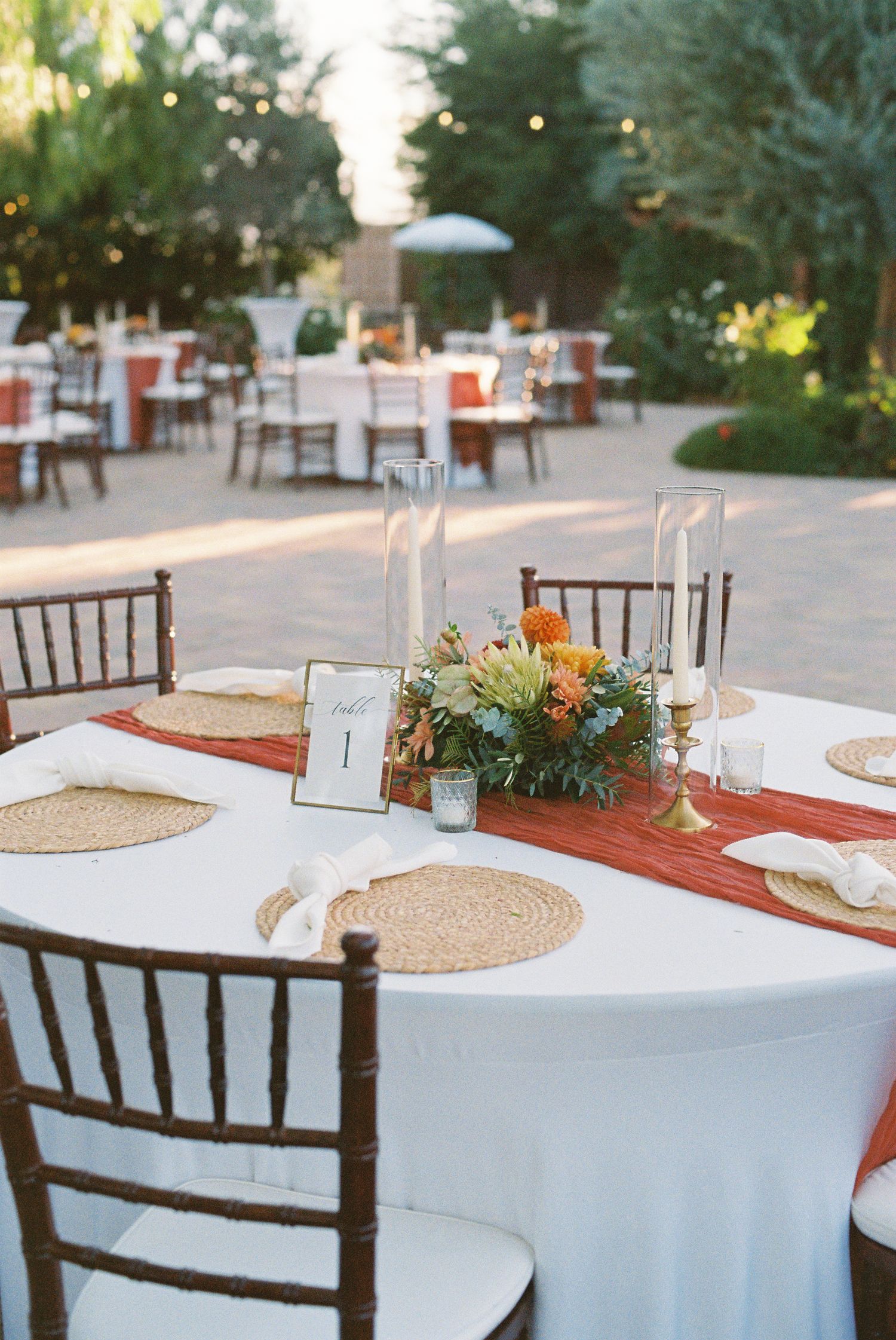 Elegant outdoor wedding reception tables with terracotta runners, floral centerpieces, and Chiavari chairs.