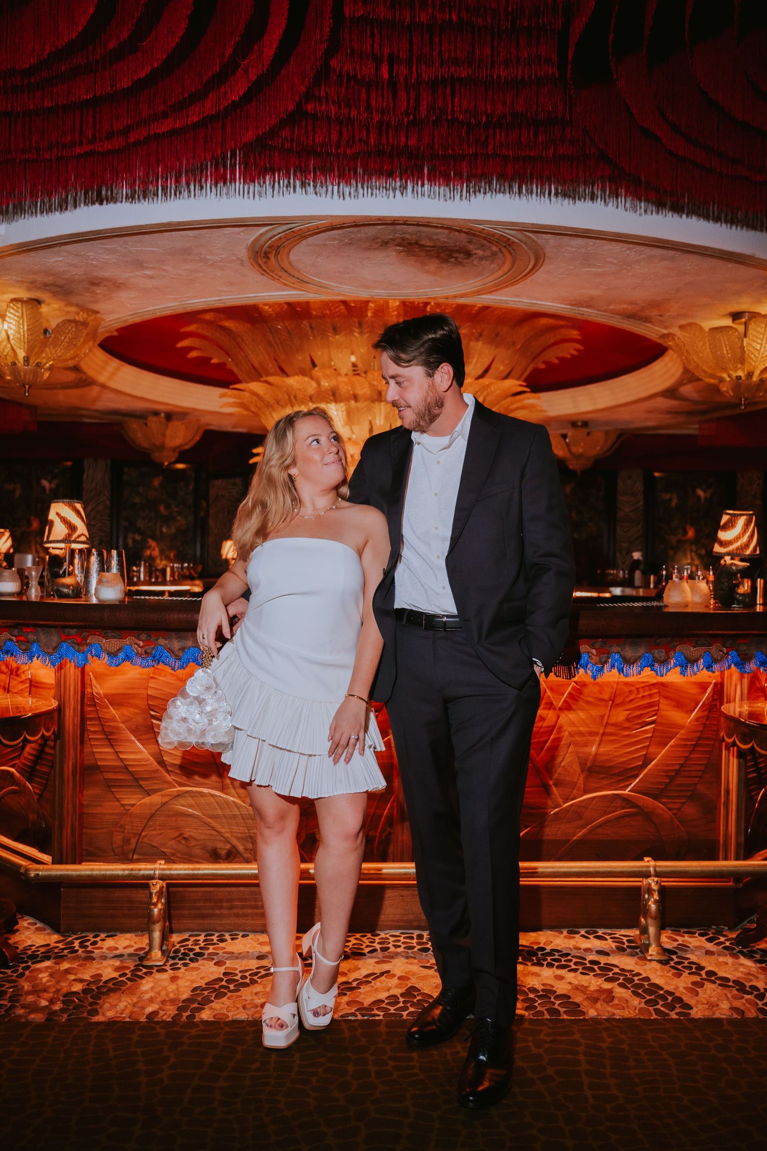 Bride and groom pose together in ornate bar venue, she wears white mini dress, he wears dark suit.