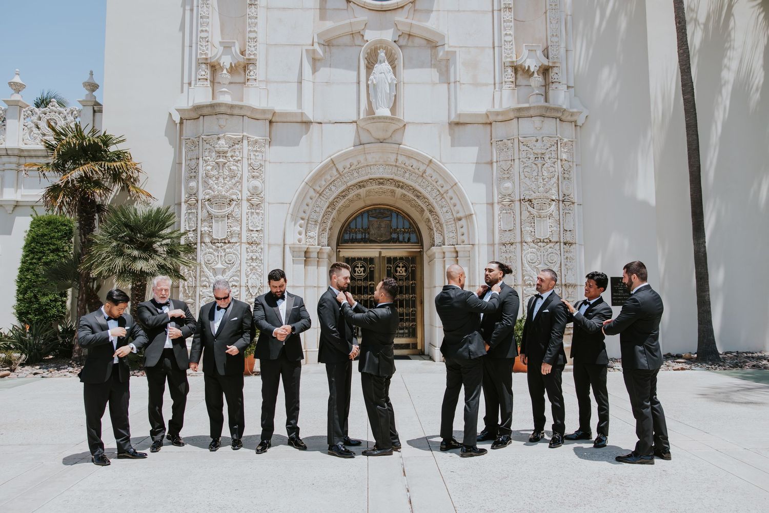Wedding party members lined up kneeling outside an ornate church facade.