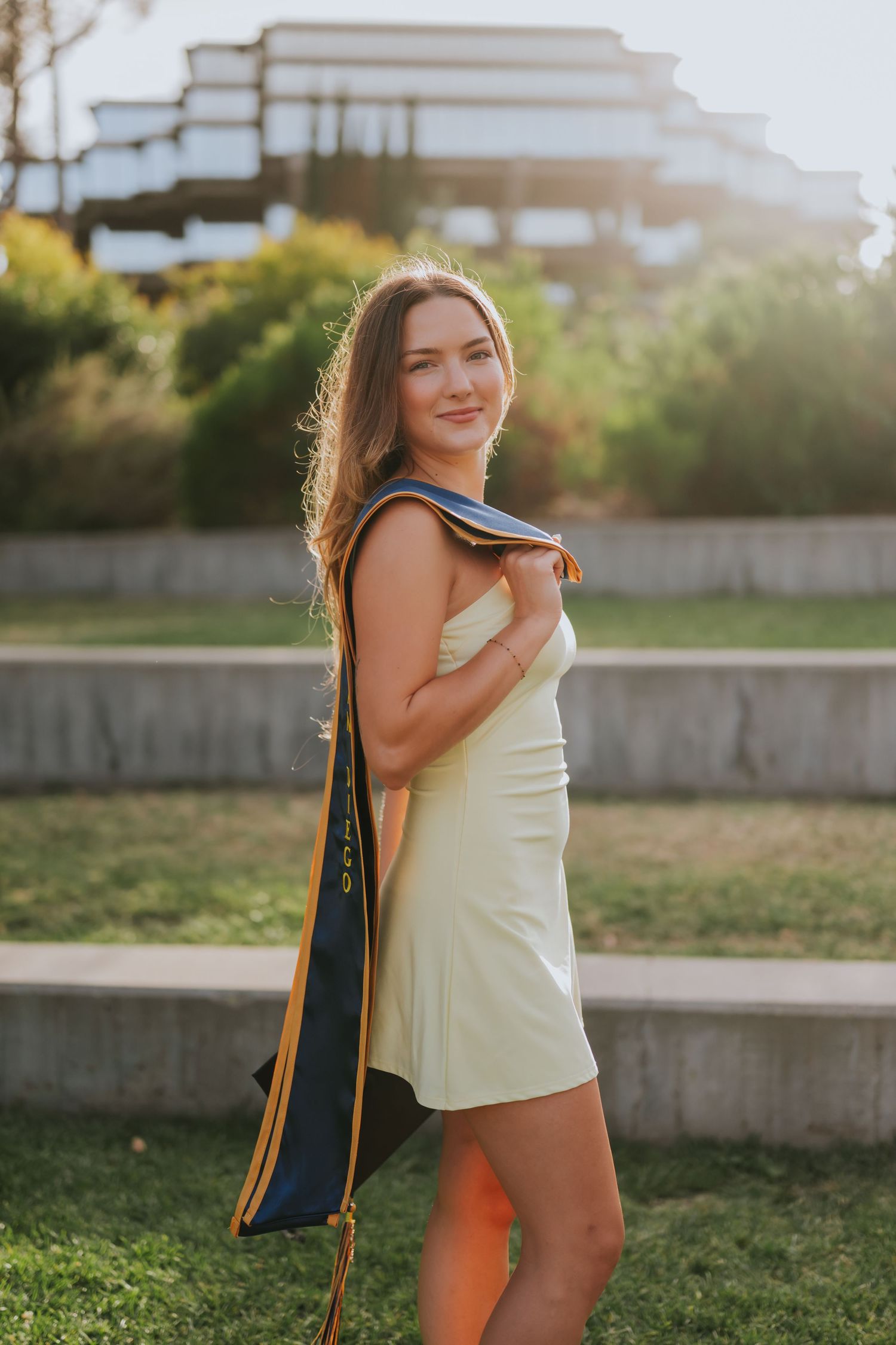 Young woman in light green mini dress carrying a yoga mat bag, smiling outdoors in golden hour sunlight.