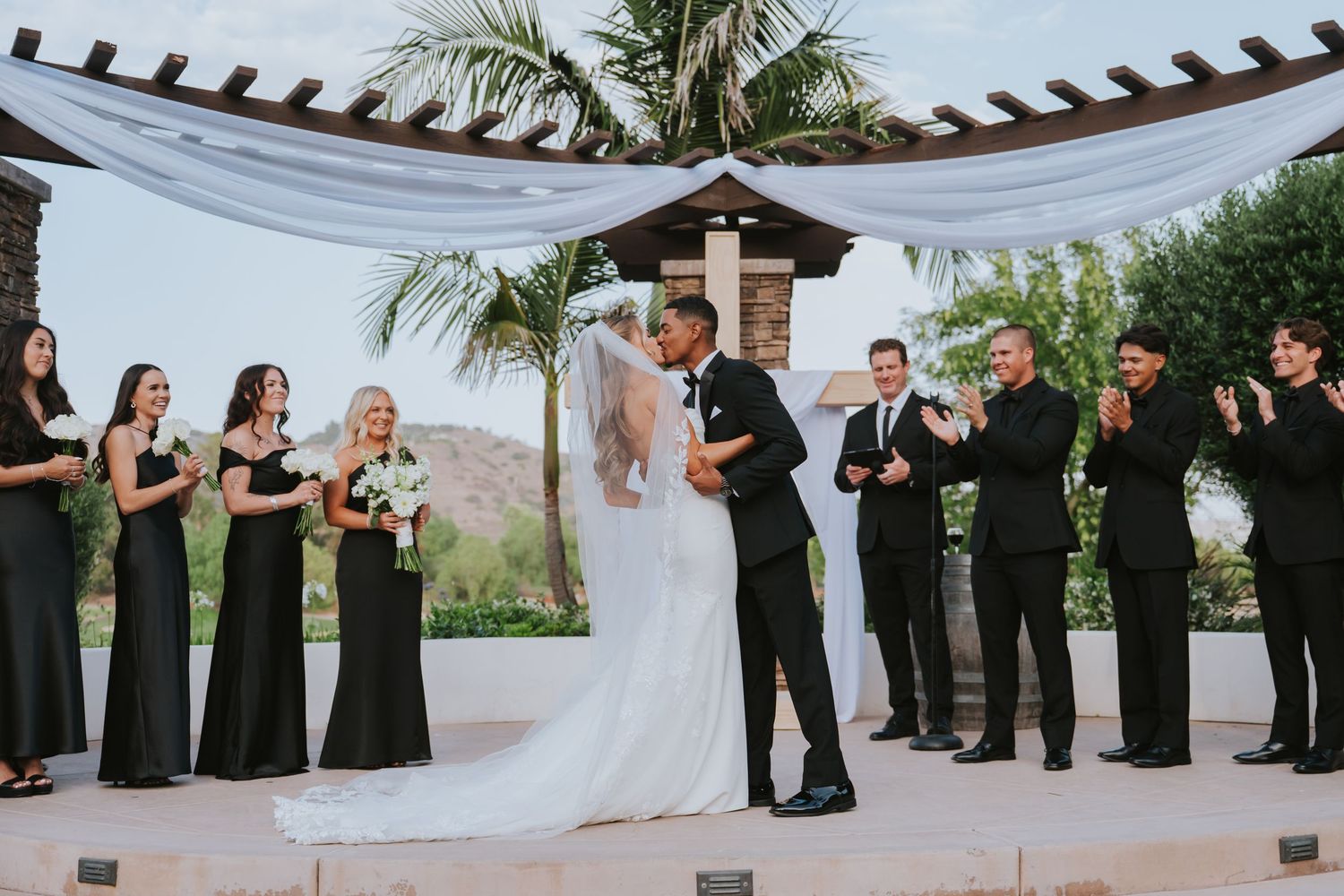 Bride and groom share first kiss at outdoor wedding ceremony with bridesmaids in black dresses and groomsmen applauding.