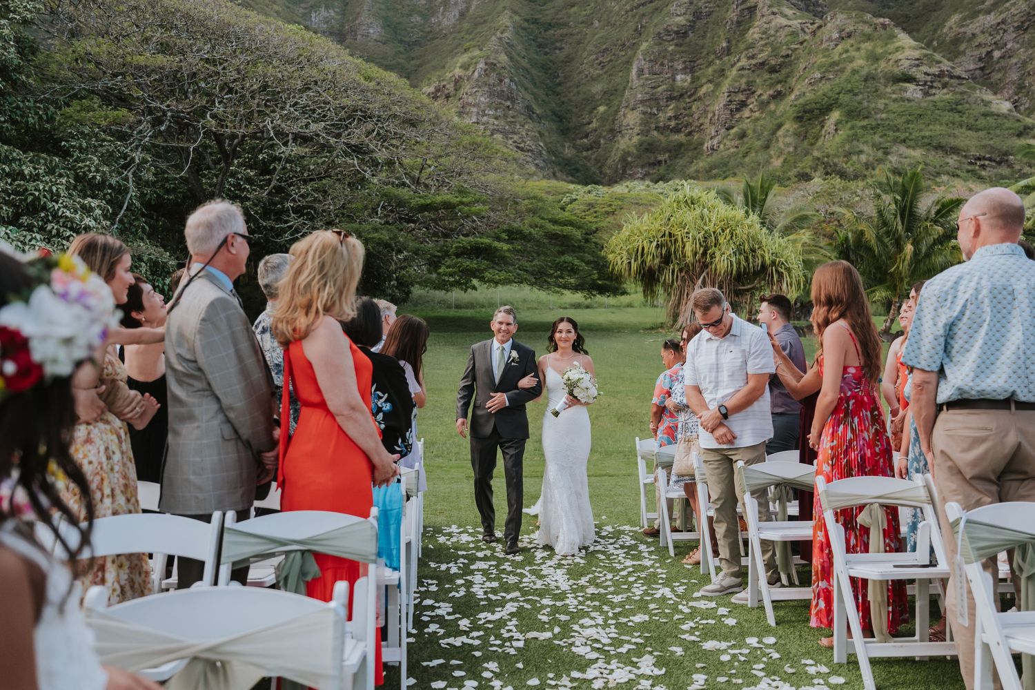 Wedding guests seated on white chairs line a grassy hillside path decorated with scattered white flower petals.