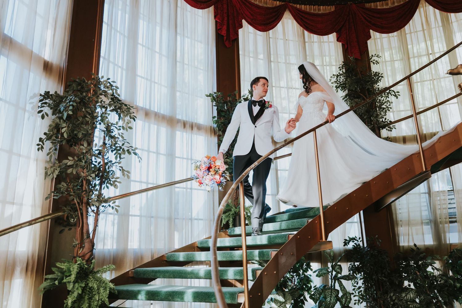 A couple in wedding attire poses elegantly on a grand staircase decorated with greenery and draped fabric.