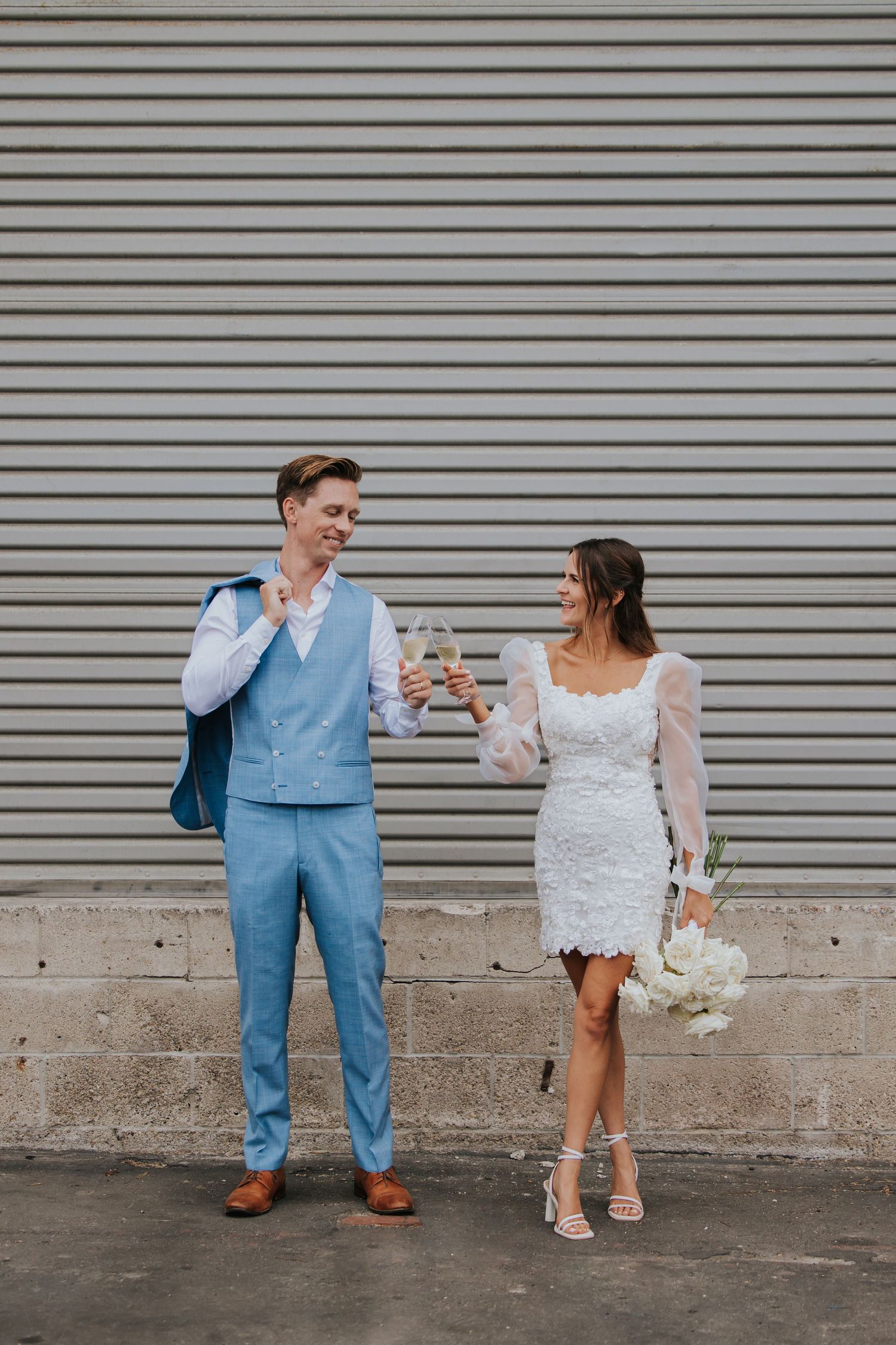 A couple's feet shown against an industrial wall, with the groom in blue suit pants and brown shoes and bride in white lace.