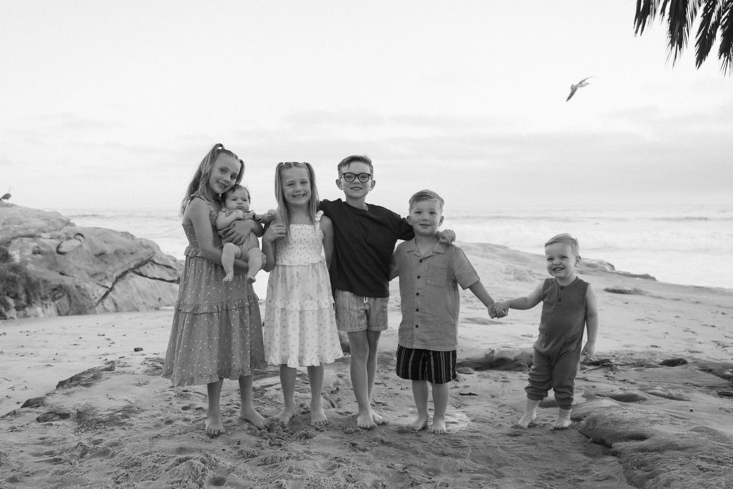 Black and white photo of five children smiling together on a sandy beach with ocean waves in the background.