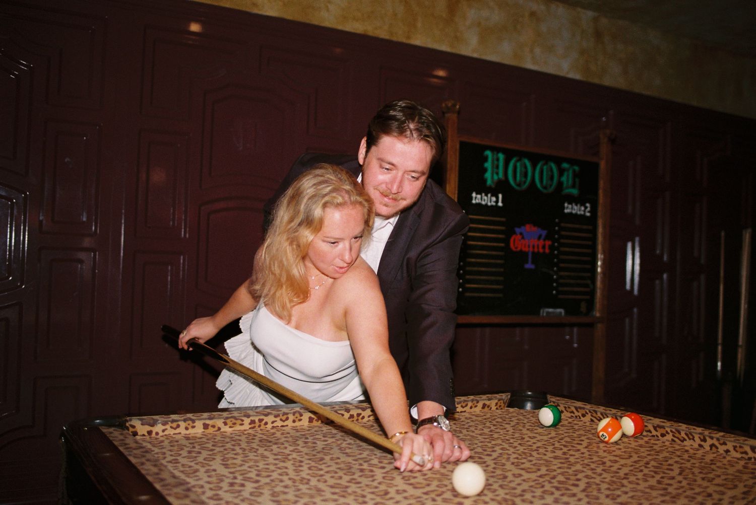 Couple playing pool together at a bar, woman in white dress leaning over billiard table with man guiding her shot.