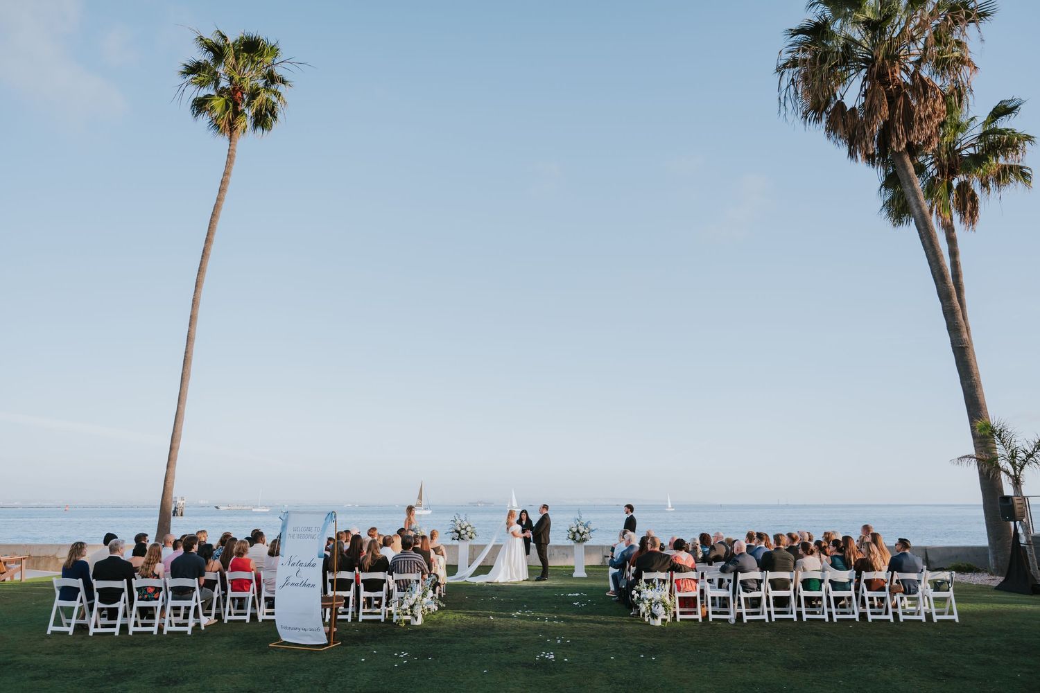 Outdoor beach wedding ceremony with guests seated on white chairs between tall palm trees overlooking the ocean.