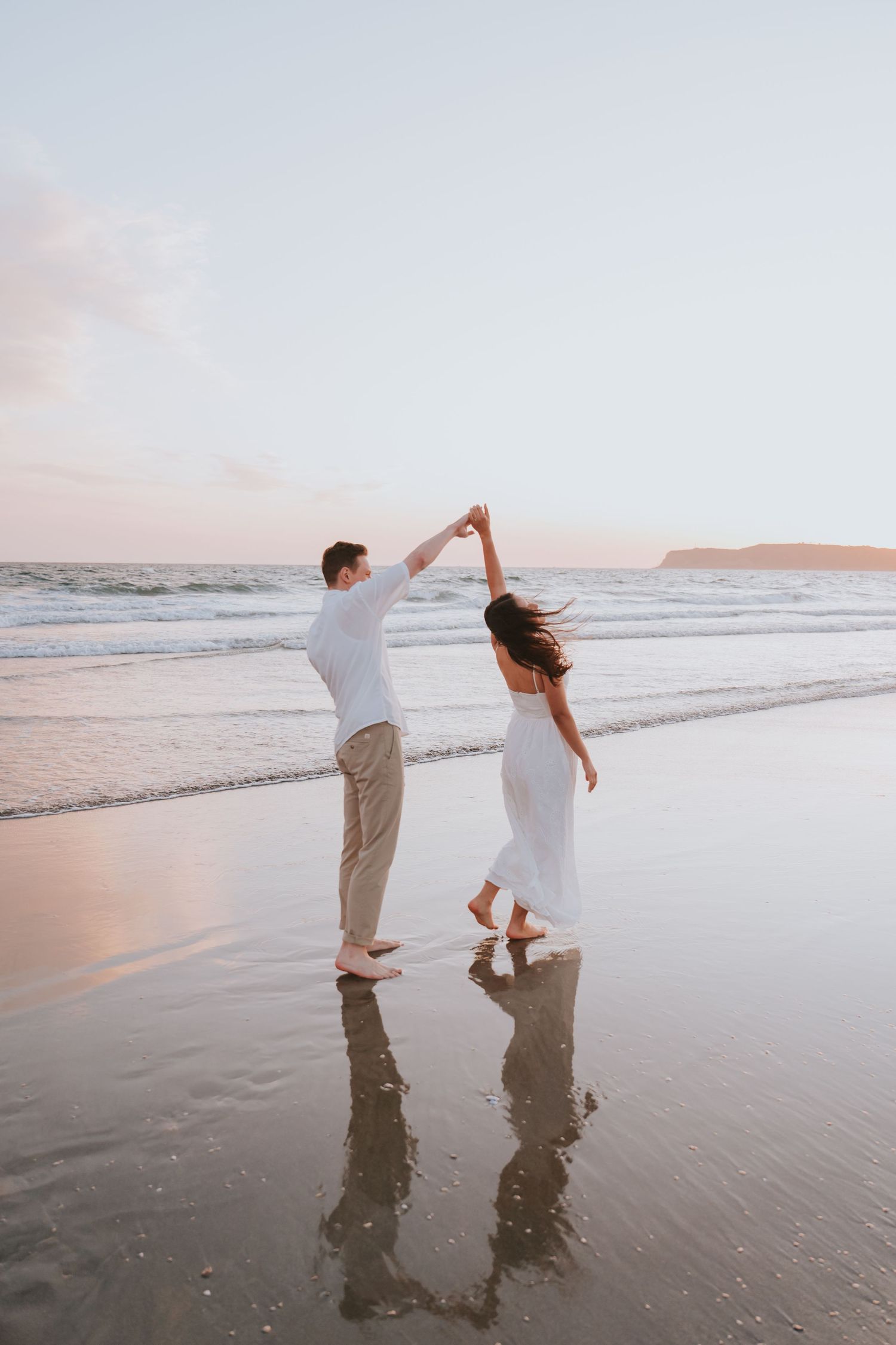 Couple dancing barefoot on a wet sandy beach at sunset, man spinning woman in white dress by ocean waves.