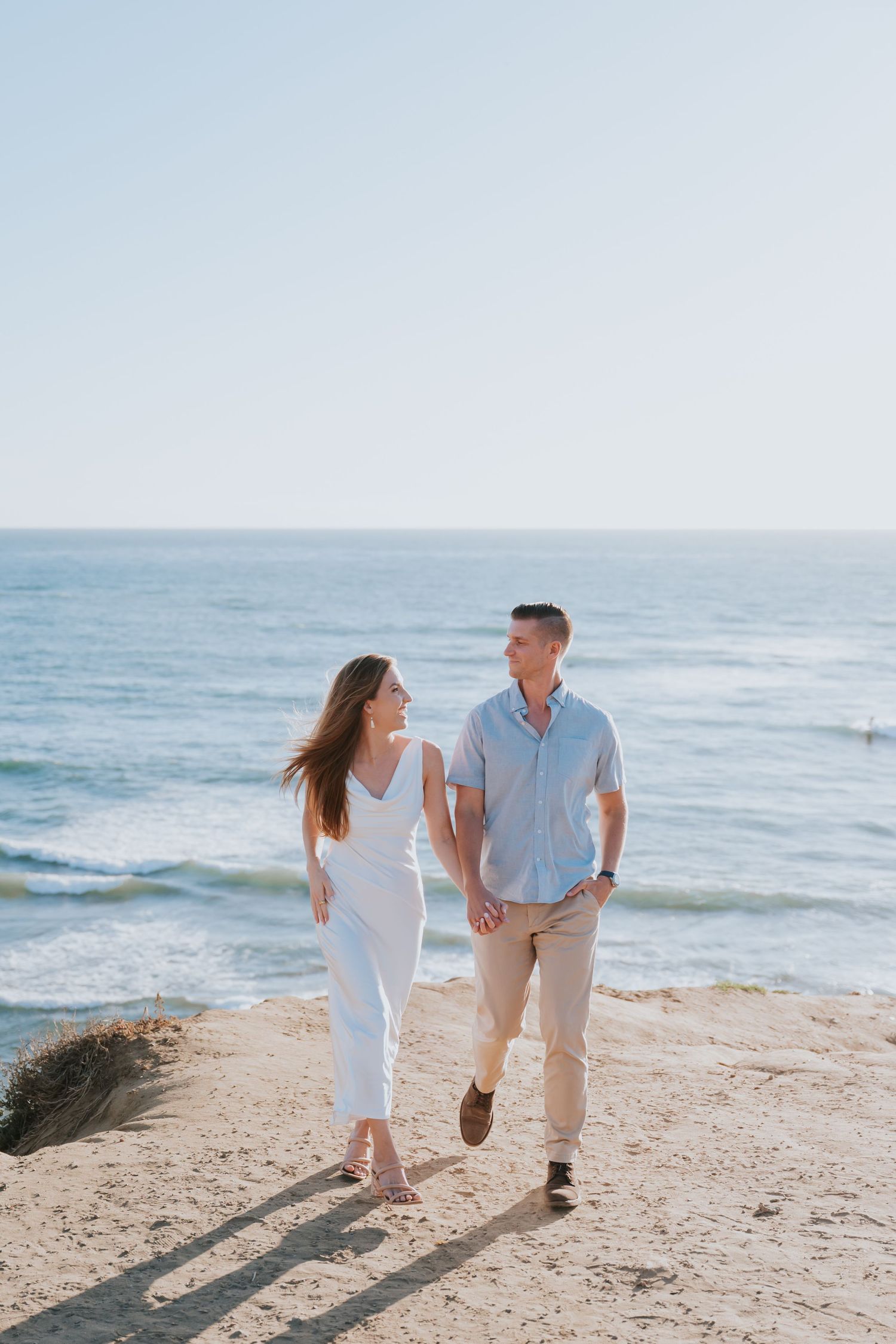 A couple walks hand-in-hand on a sunny beach, smiling at each other with the ocean in the background.