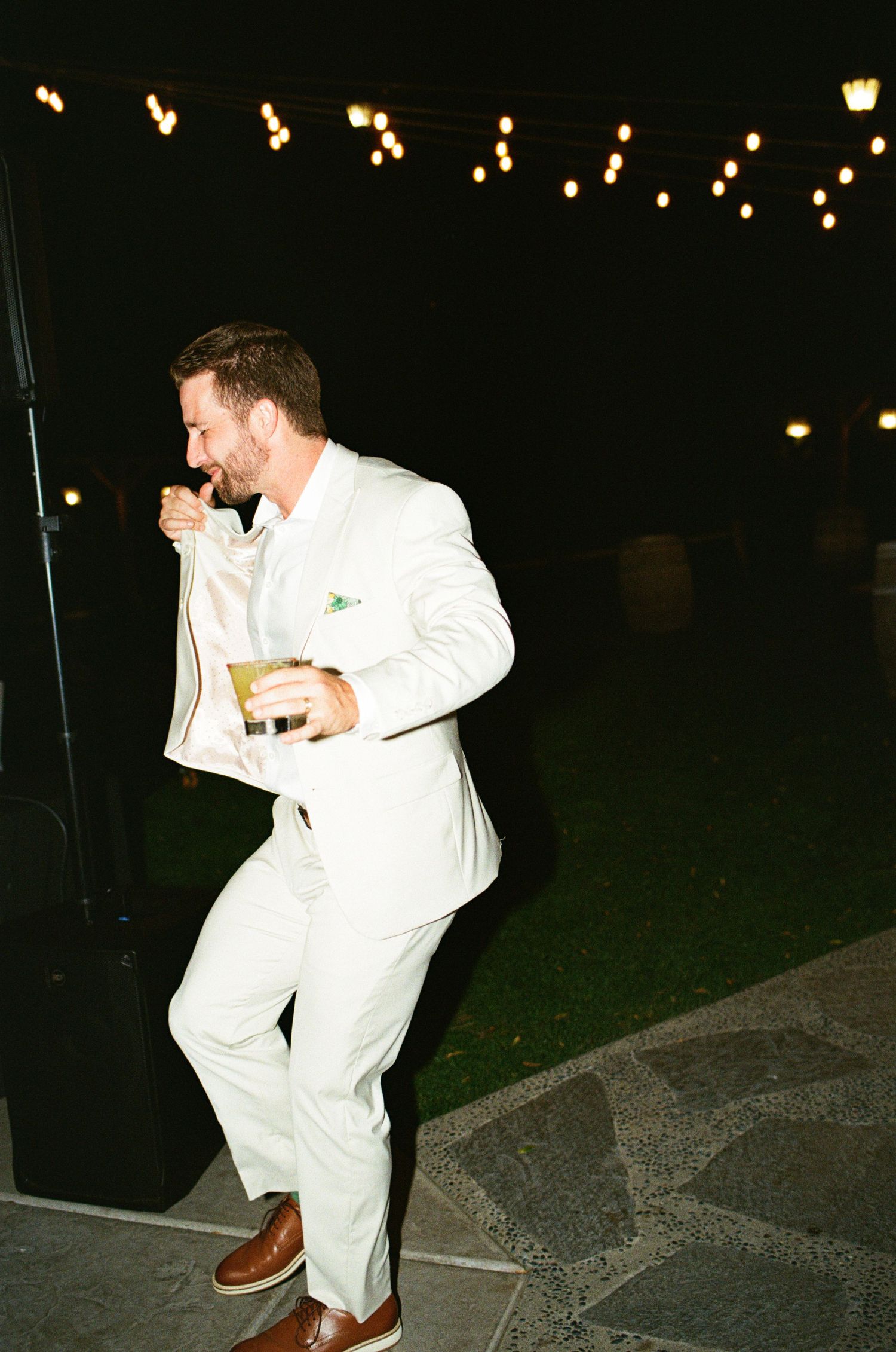 Groom in white suit holds drink under string lights at outdoor wedding reception at night.