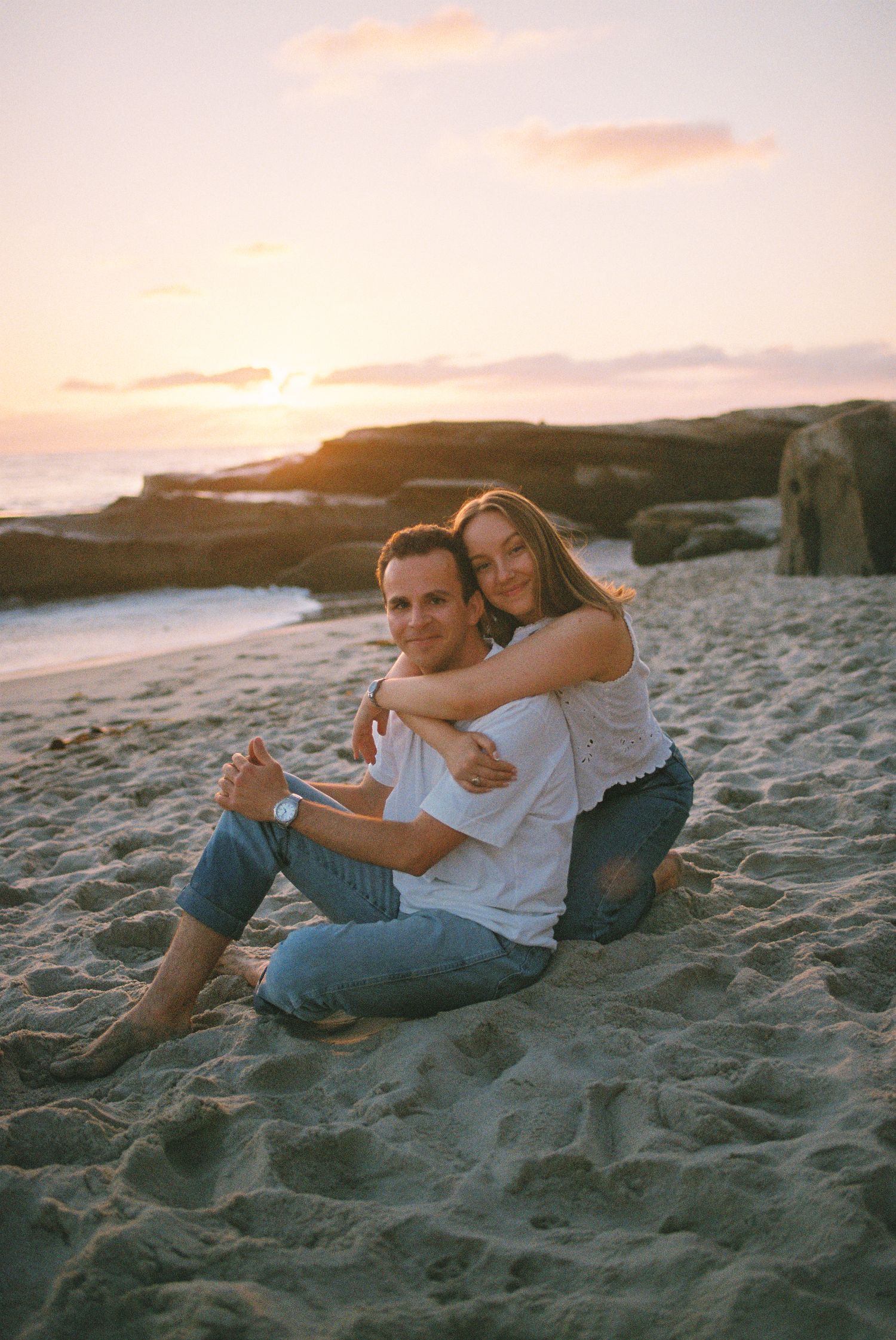 Couple sitting on sandy beach at sunset, woman hugging man from behind, both wearing white tops and jeans.