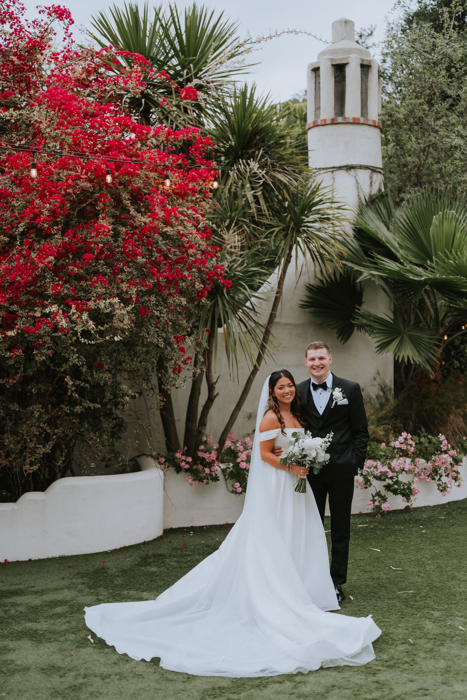 Wedding portrait in a Spanish garden with vibrant red bougainvillea and white tower.