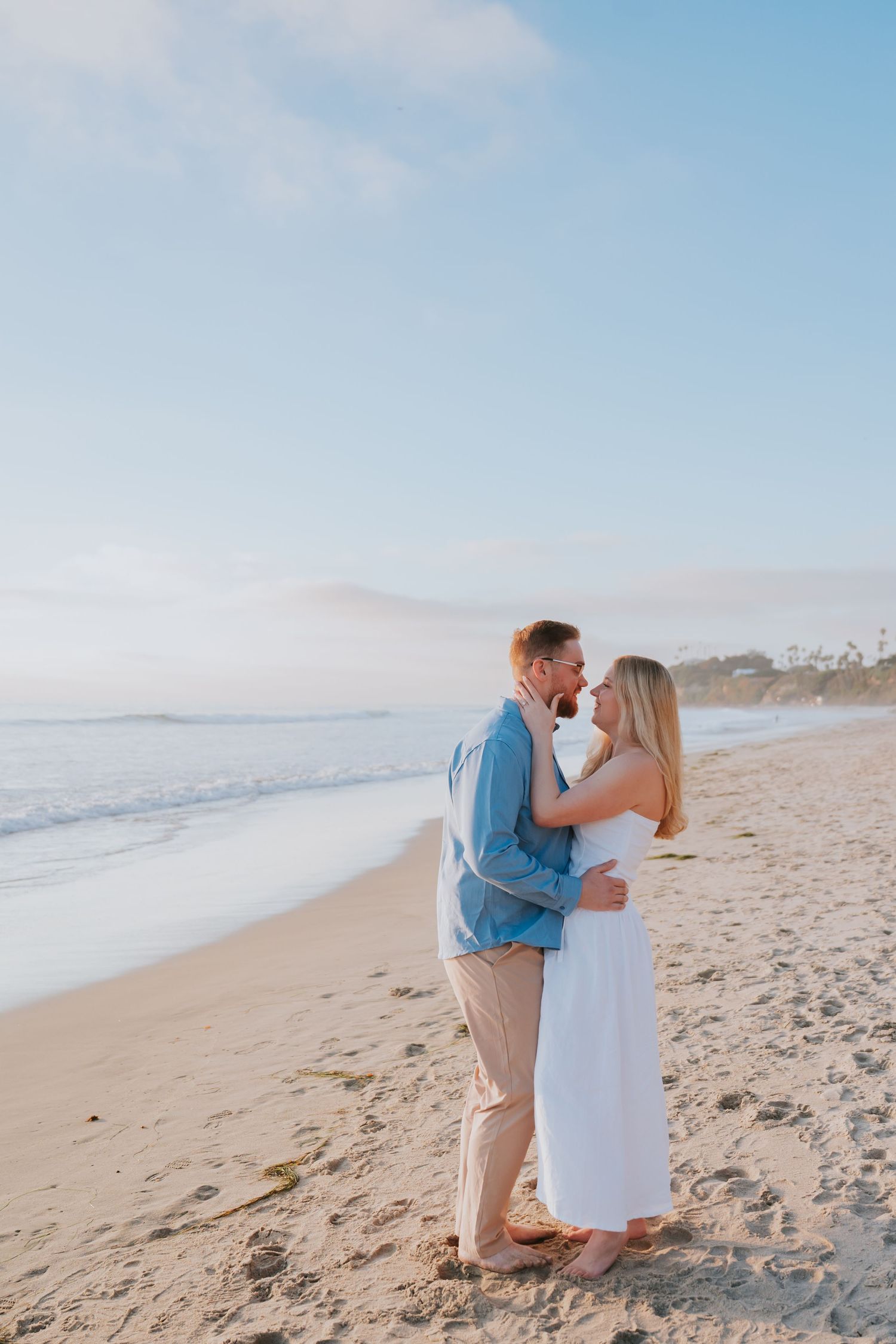 Couple sharing a romantic kiss on a sandy beach at sunset, man in blue shirt, woman in white dress.