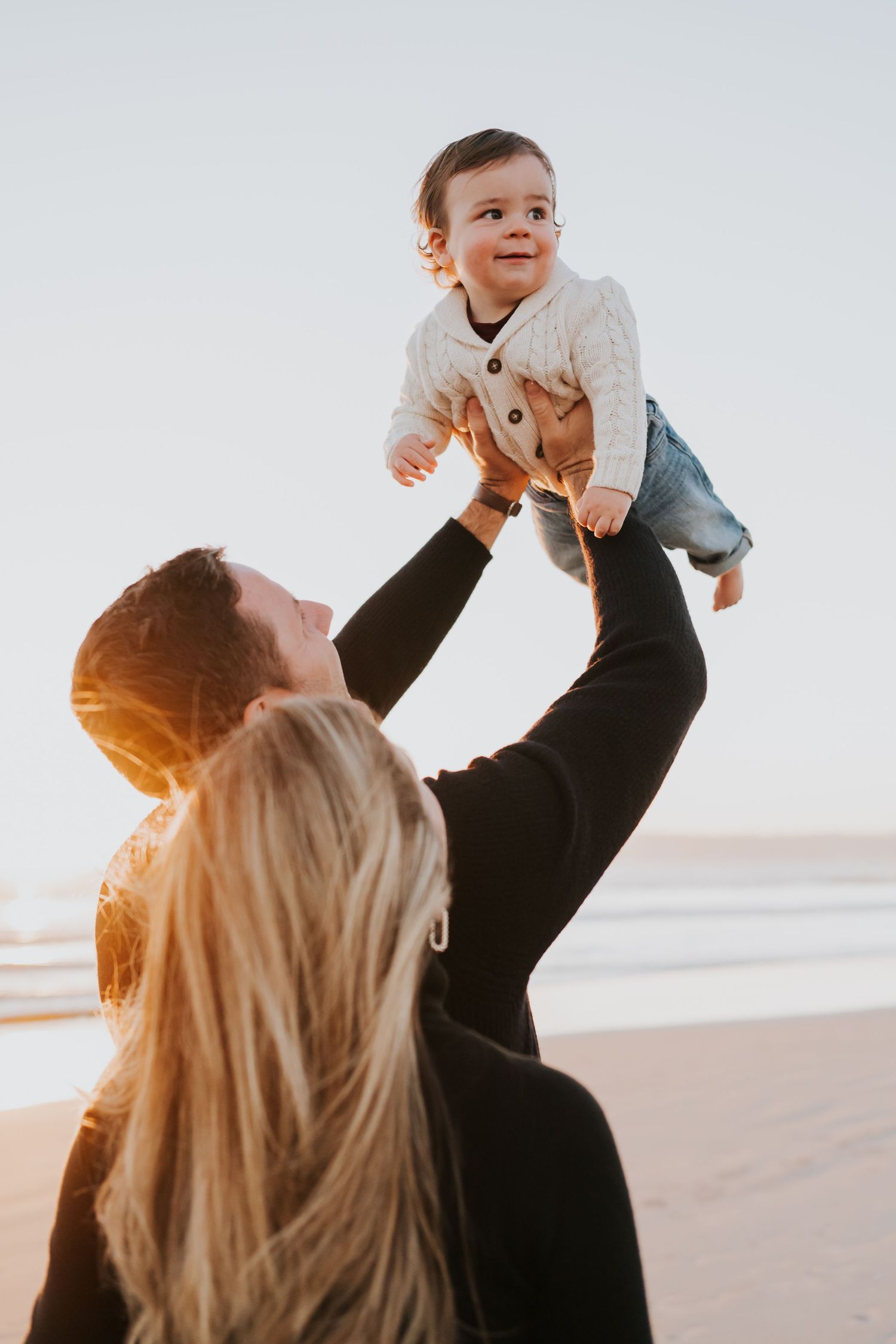 Parents lifting a happy baby in the air on a sunny beach at golden hour.