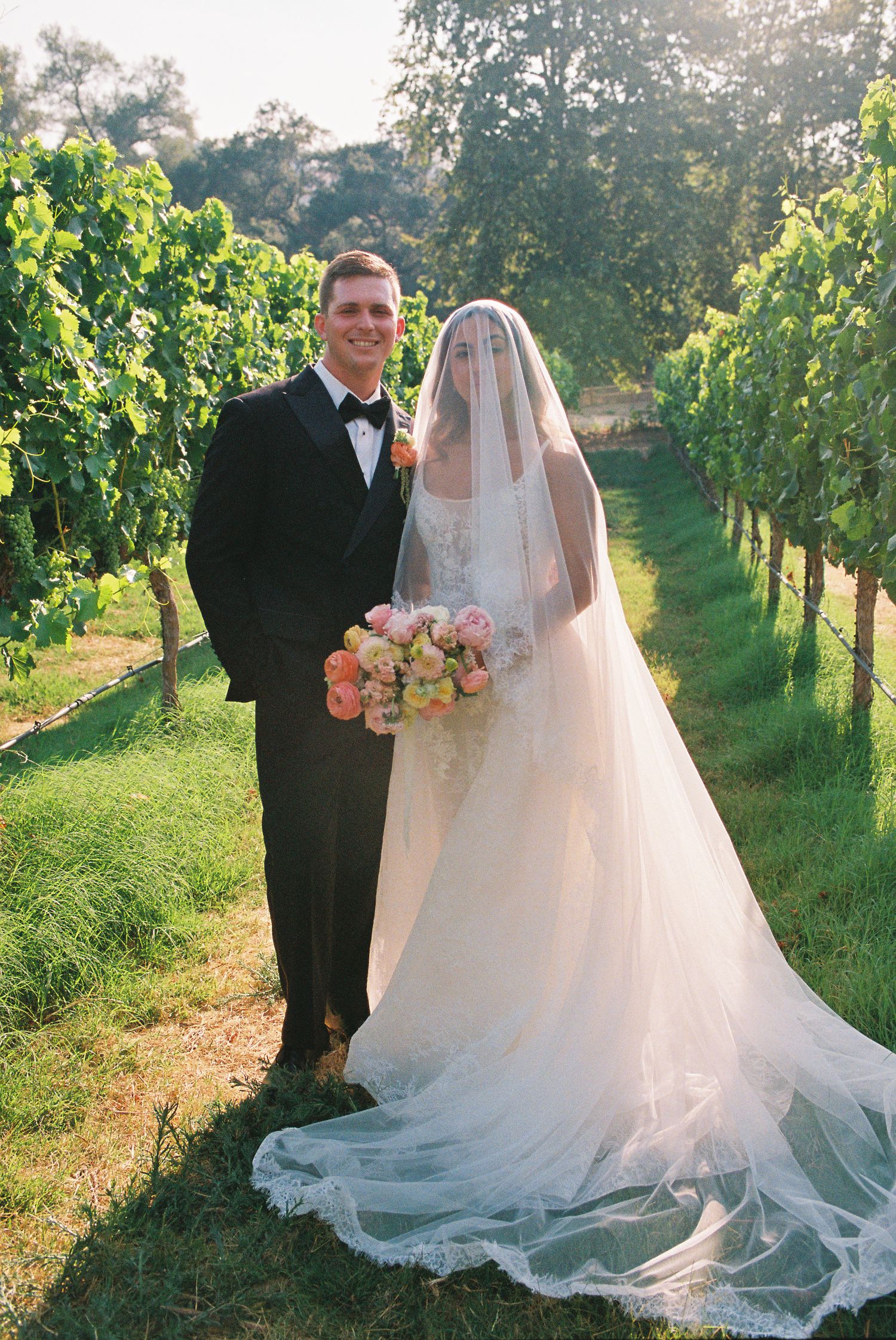 Bride and groom pose together in a sunlit vineyard, bride wearing a long veil and gown holding a pink bouquet.