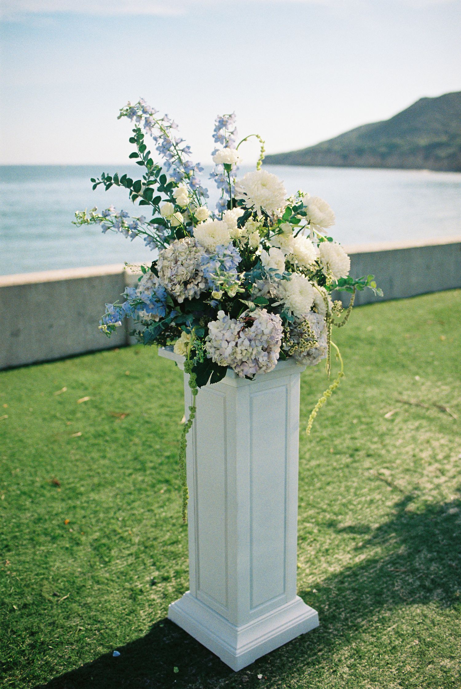 White pedestal with blue and white floral arrangement overlooking ocean and mountains at outdoor wedding ceremony.