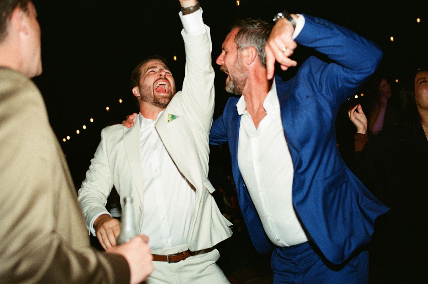 Two men in suits dancing joyfully at a wedding reception, one in light gray and one in royal blue.
