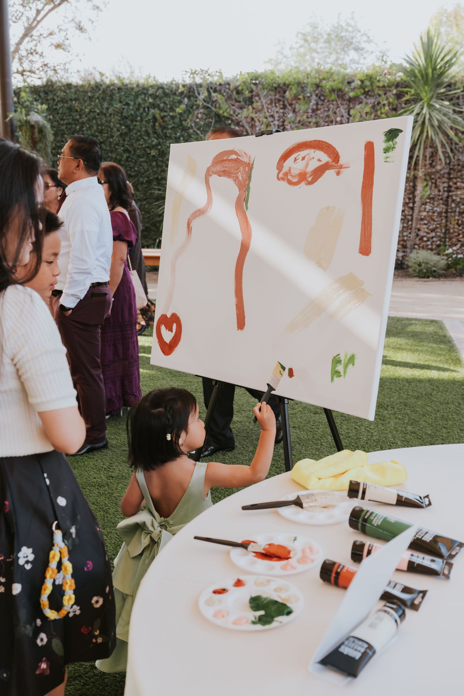 Wedding guests participate in a creative painting activity with art supplies laid out on outdoor tables.