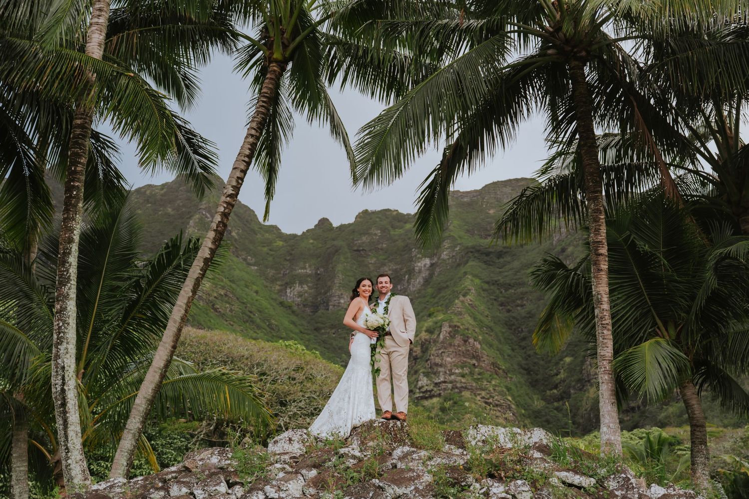 Newlyweds pose for portrait with majestic Hawaiian mountains and palm trees in background.