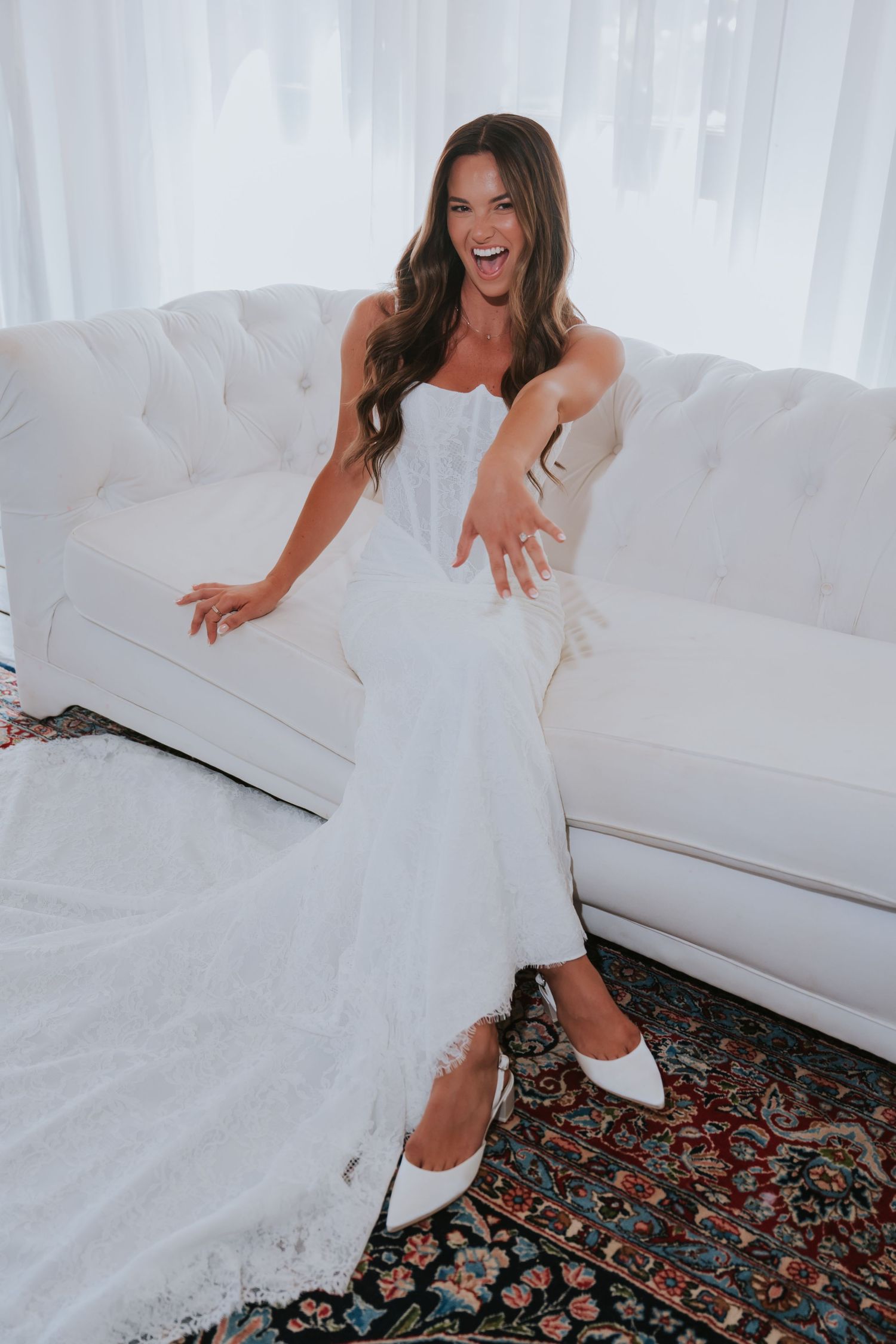 Smiling bride in white strapless wedding gown and heels sitting on tufted white sofa with colorful rug.