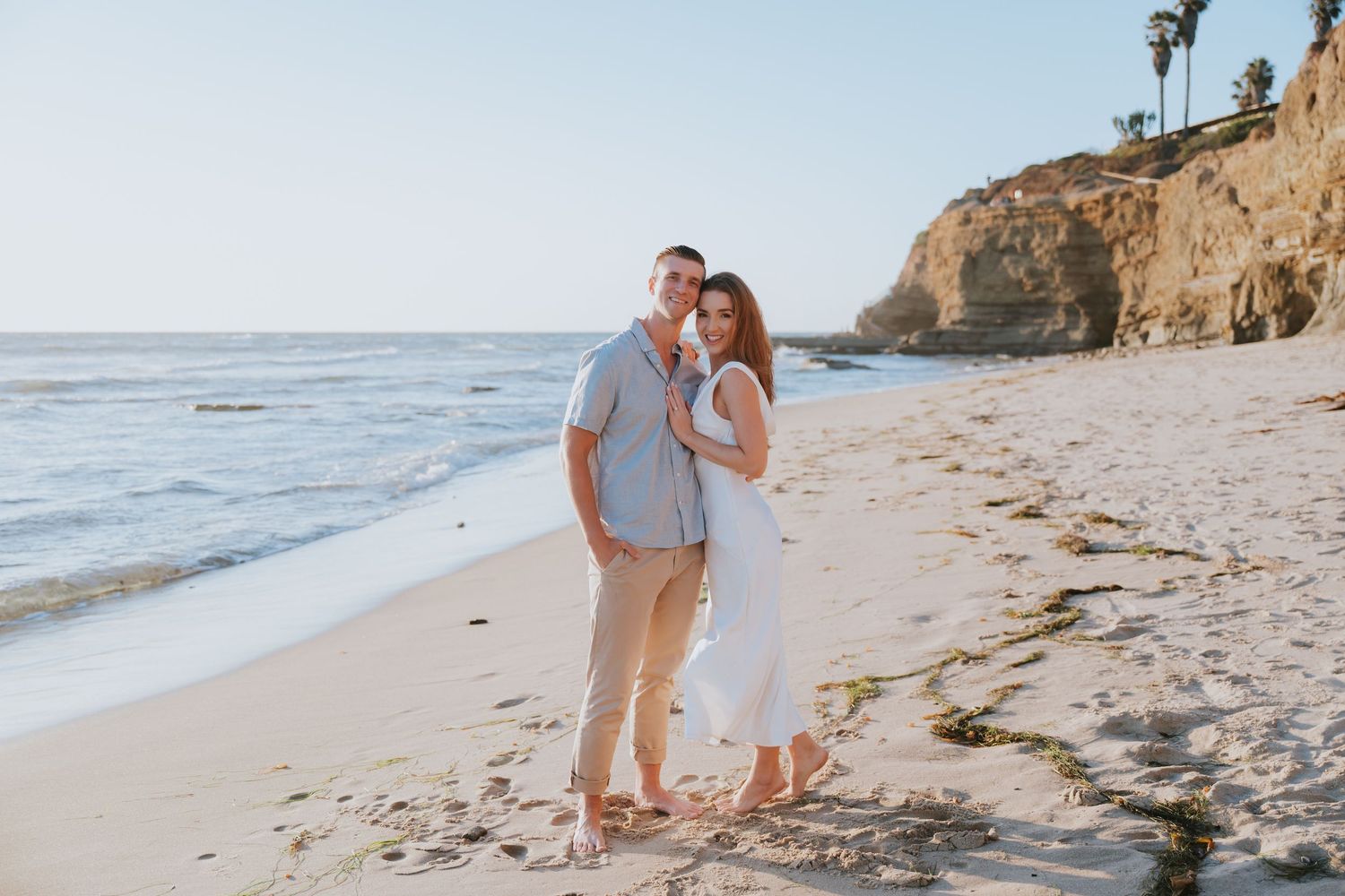 Couple embracing and smiling on a sandy beach with ocean waves and rocky cliffs in the background.