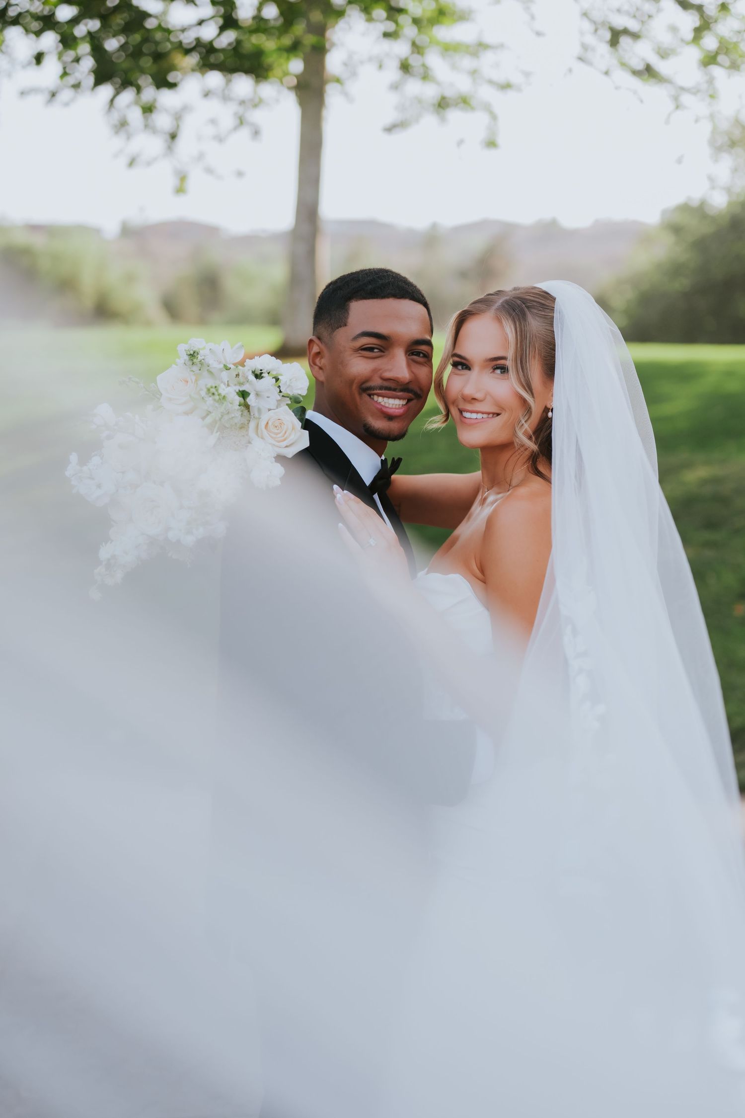 Interracial couple in wedding attire smiling outdoors, groom in tuxedo, bride in white gown with veil holding bouquet.