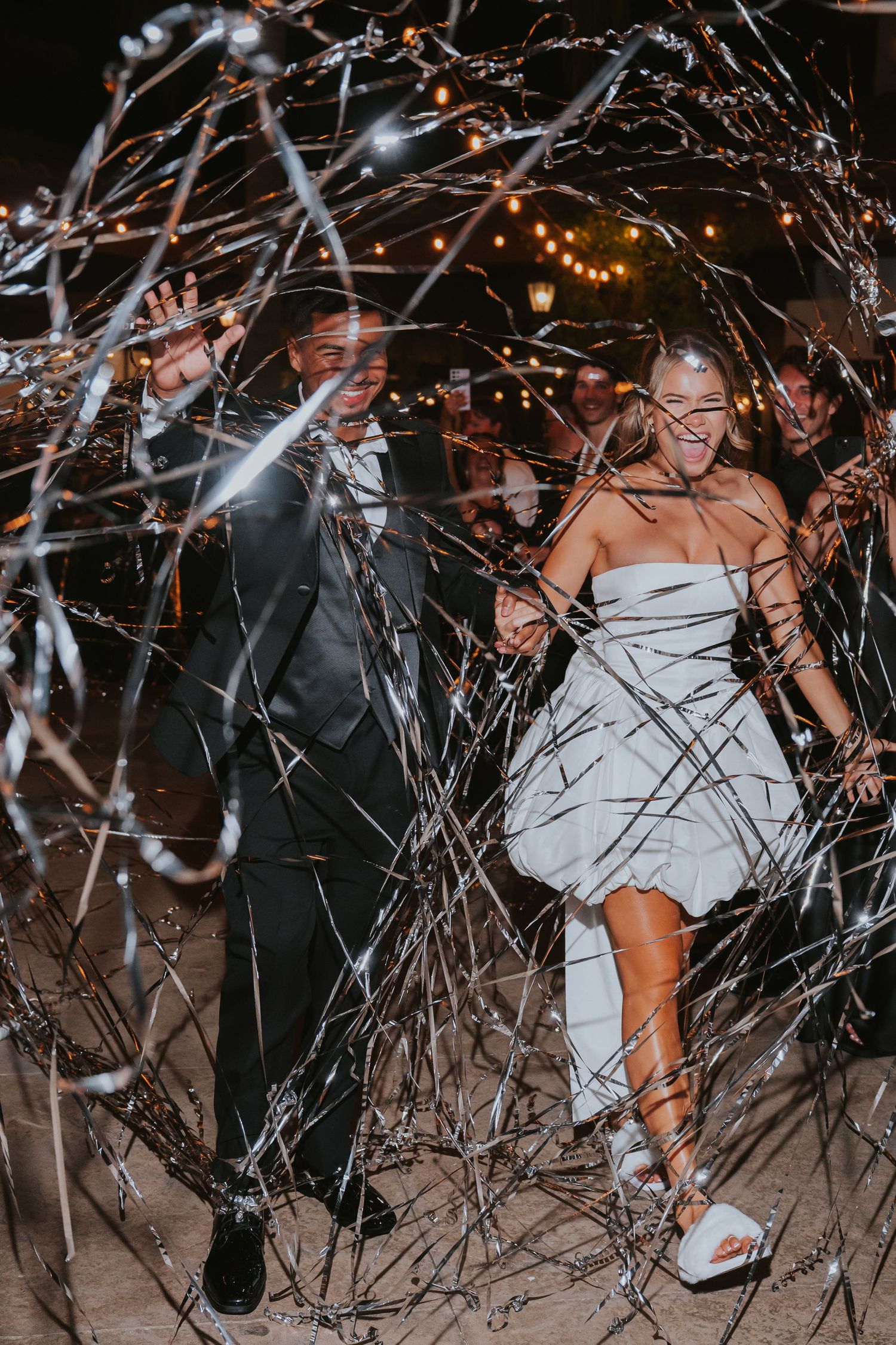 Bride and groom exit through sparklers at night, bride in short white dress, groom in dark suit, bokeh lights behind.