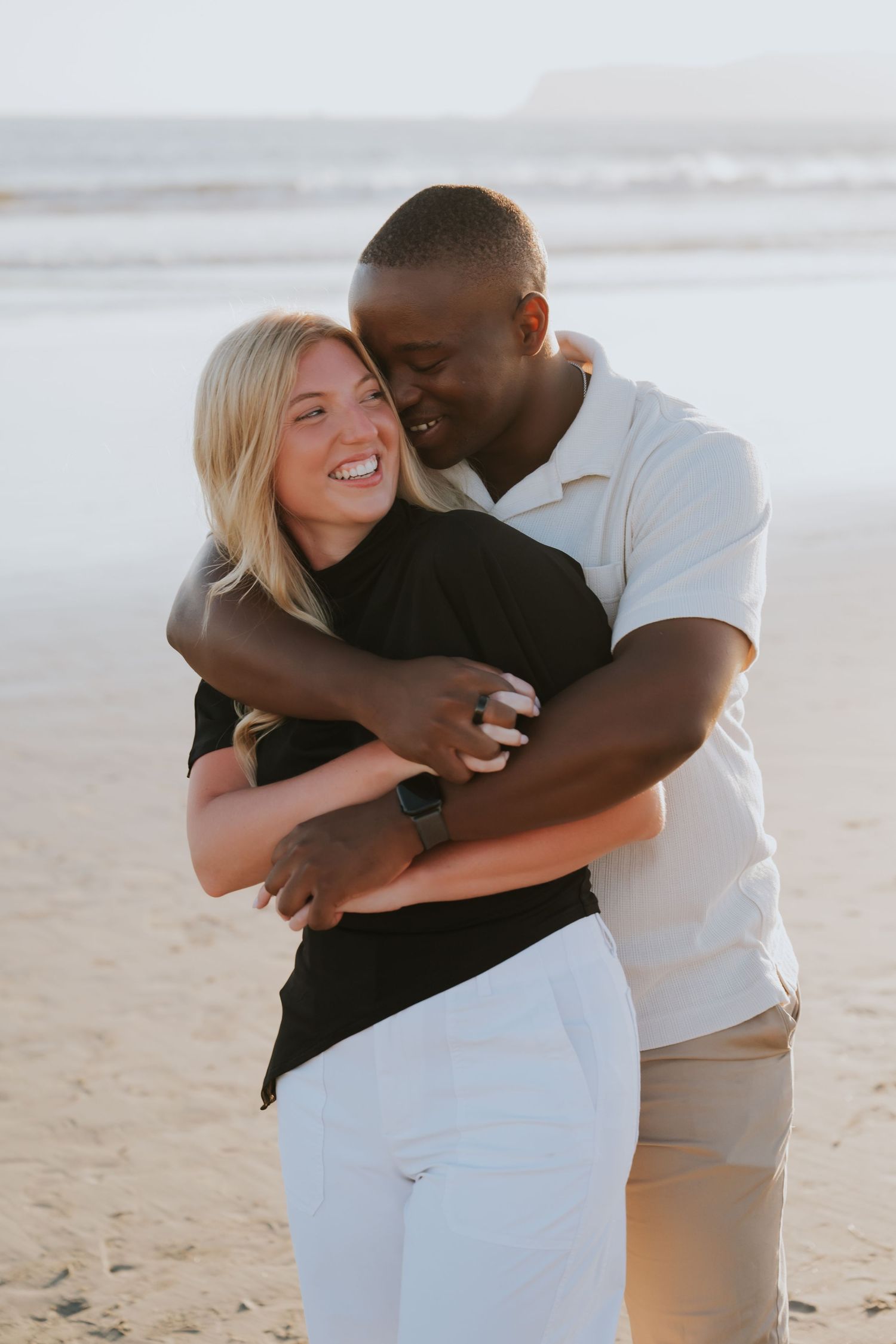 Interracial couple embracing on a sunny beach, smiling woman in black top and white pants.