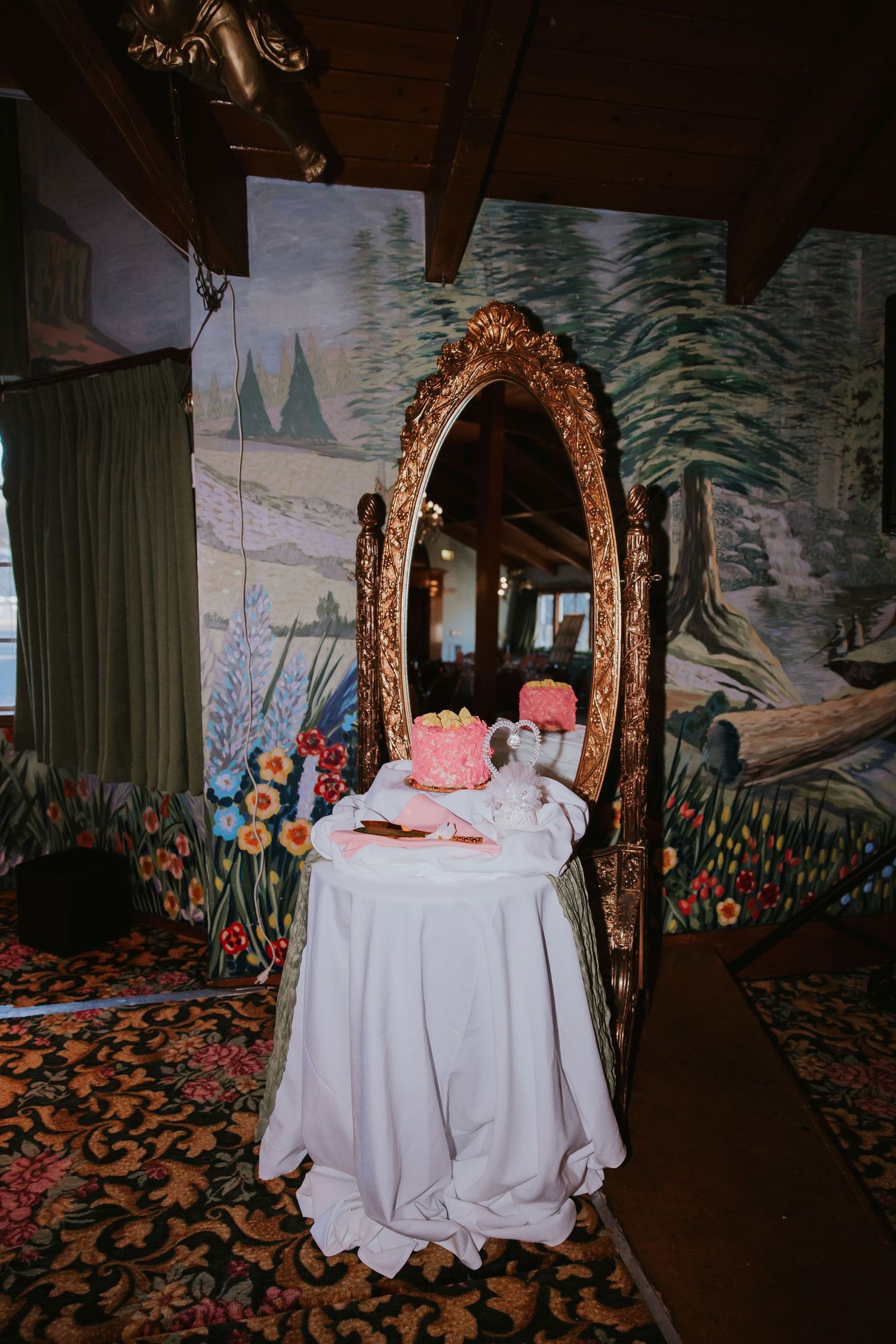 Wedding venue space with ornate gold mirrors, white tablecloths, and whimsical wall murals depicting forest scenes.