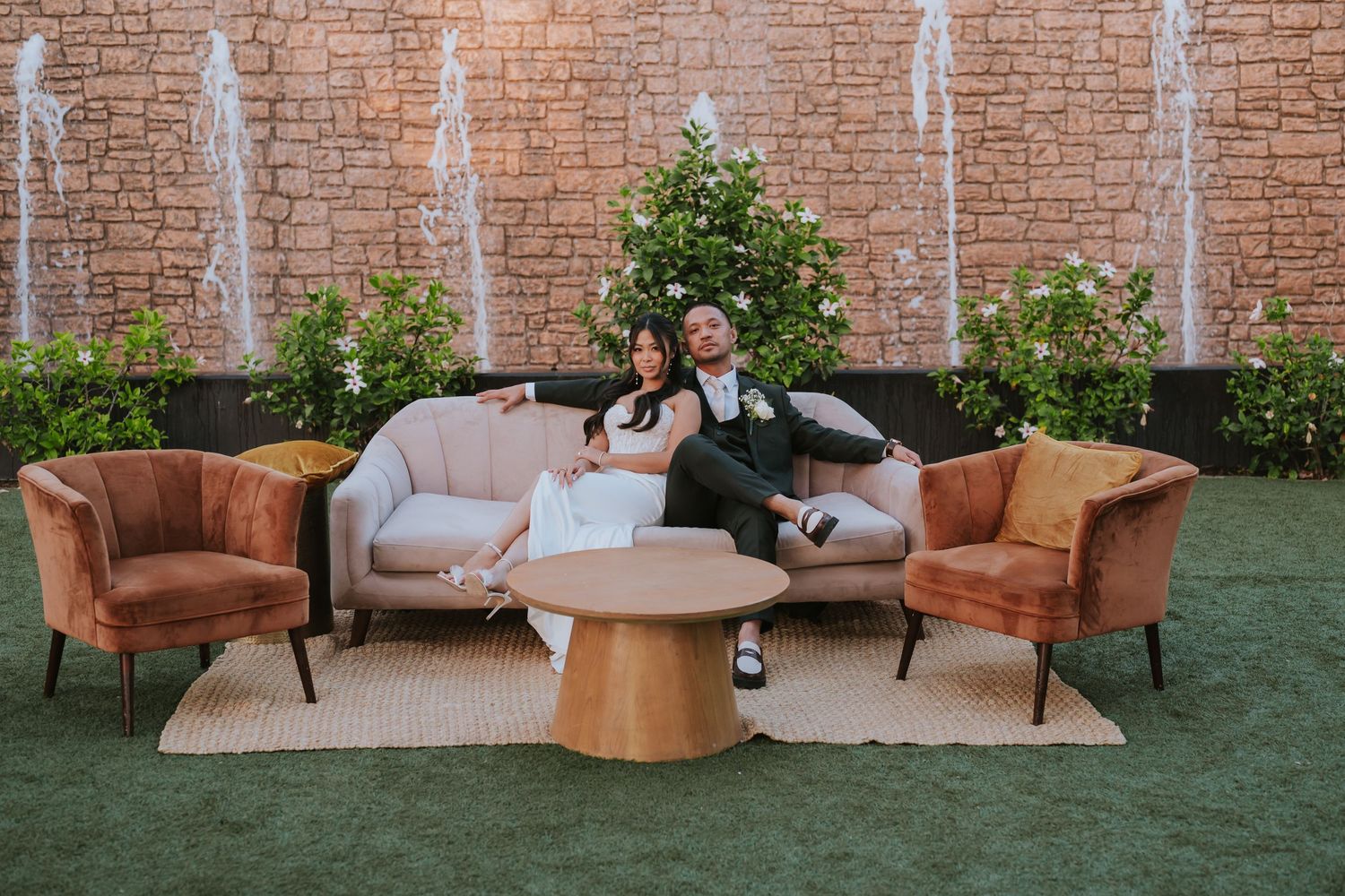 Newlyweds relax on an outdoor lounge setup with brown velvet chairs and a stone wall backdrop.