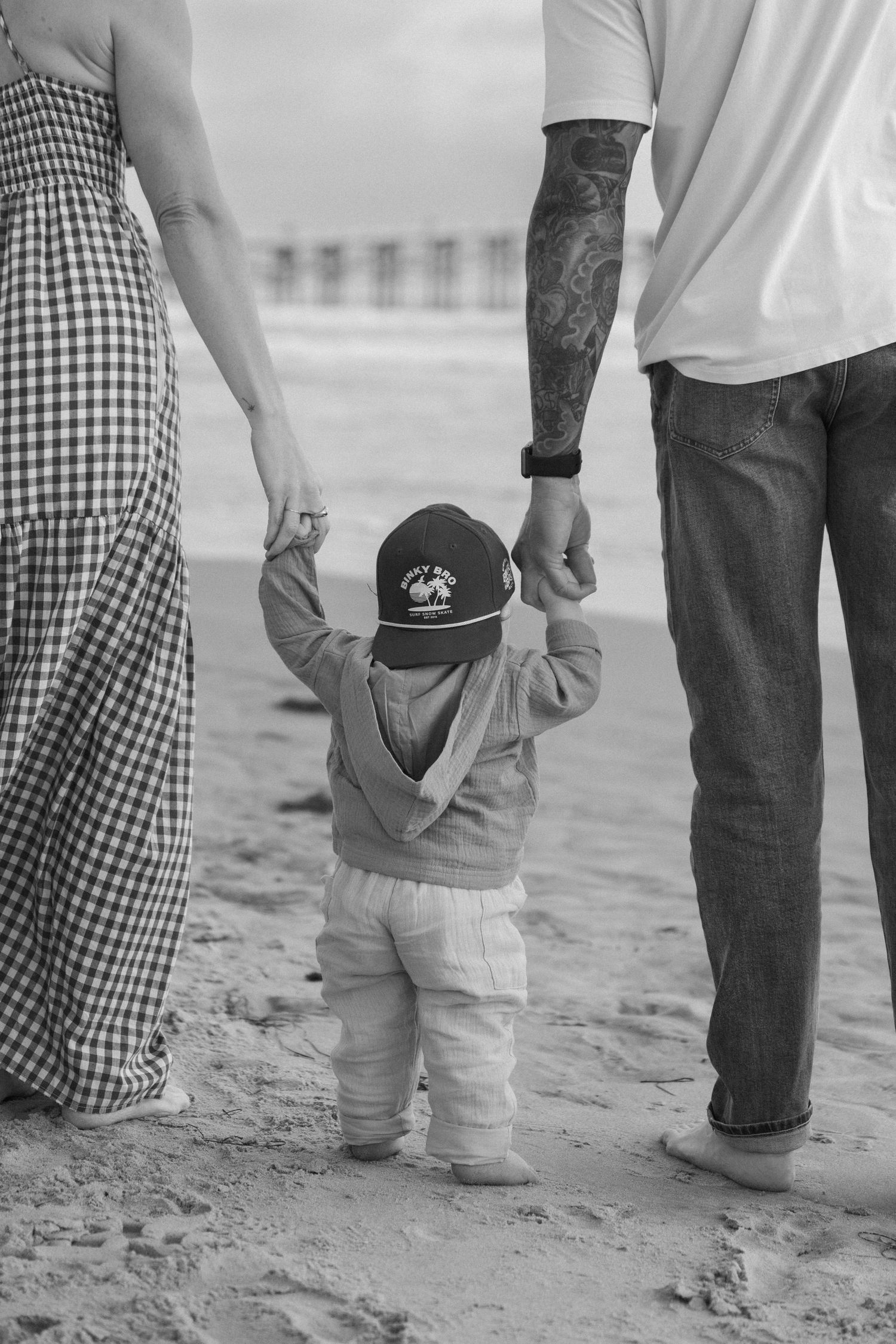 Black and white photo of a toddler holding parents hands on a sandy beach, wearing a cap and hoodie.