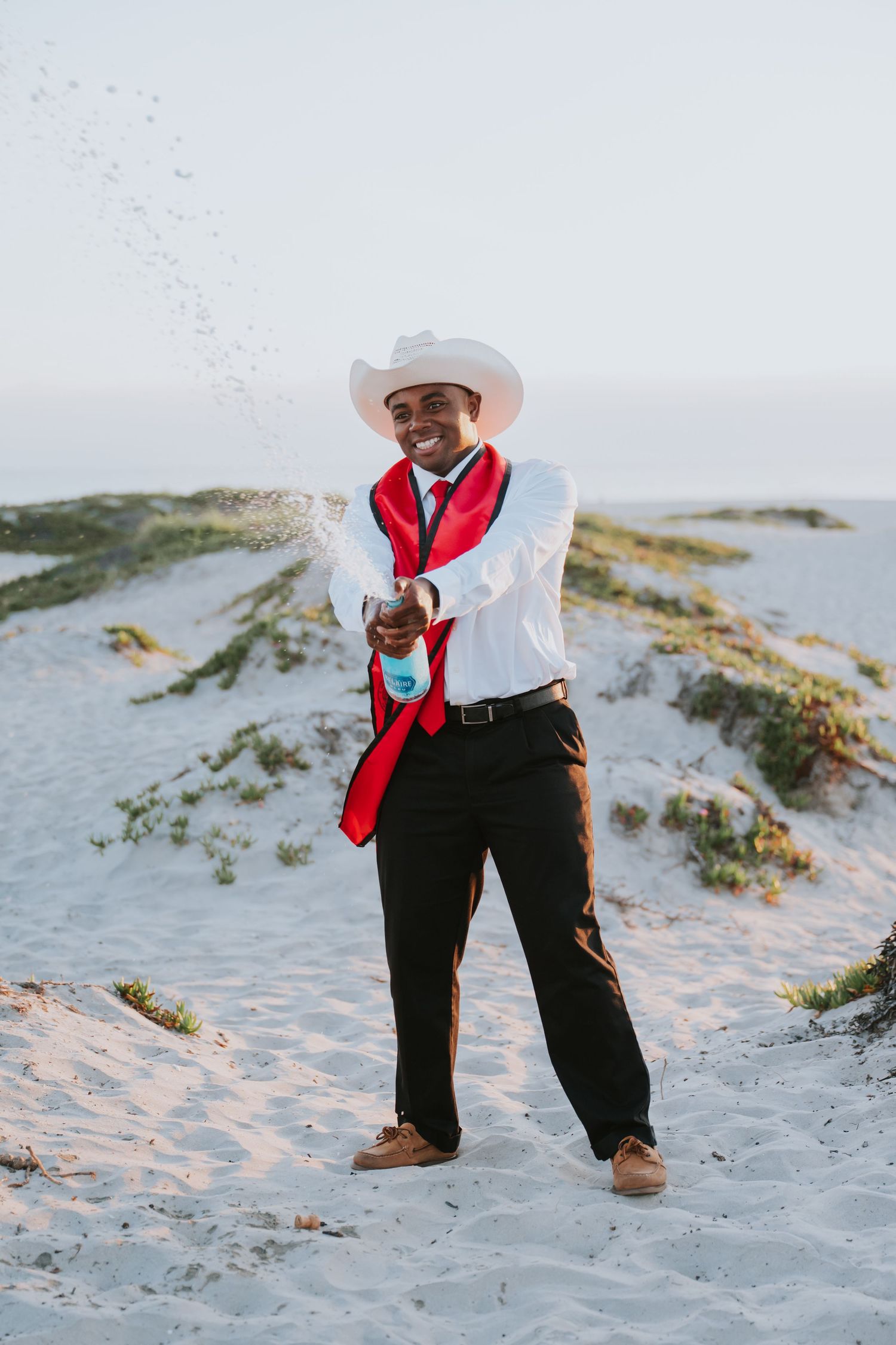 Man in white cowboy hat, red tie, and white shirt pops champagne on sandy beach dunes.