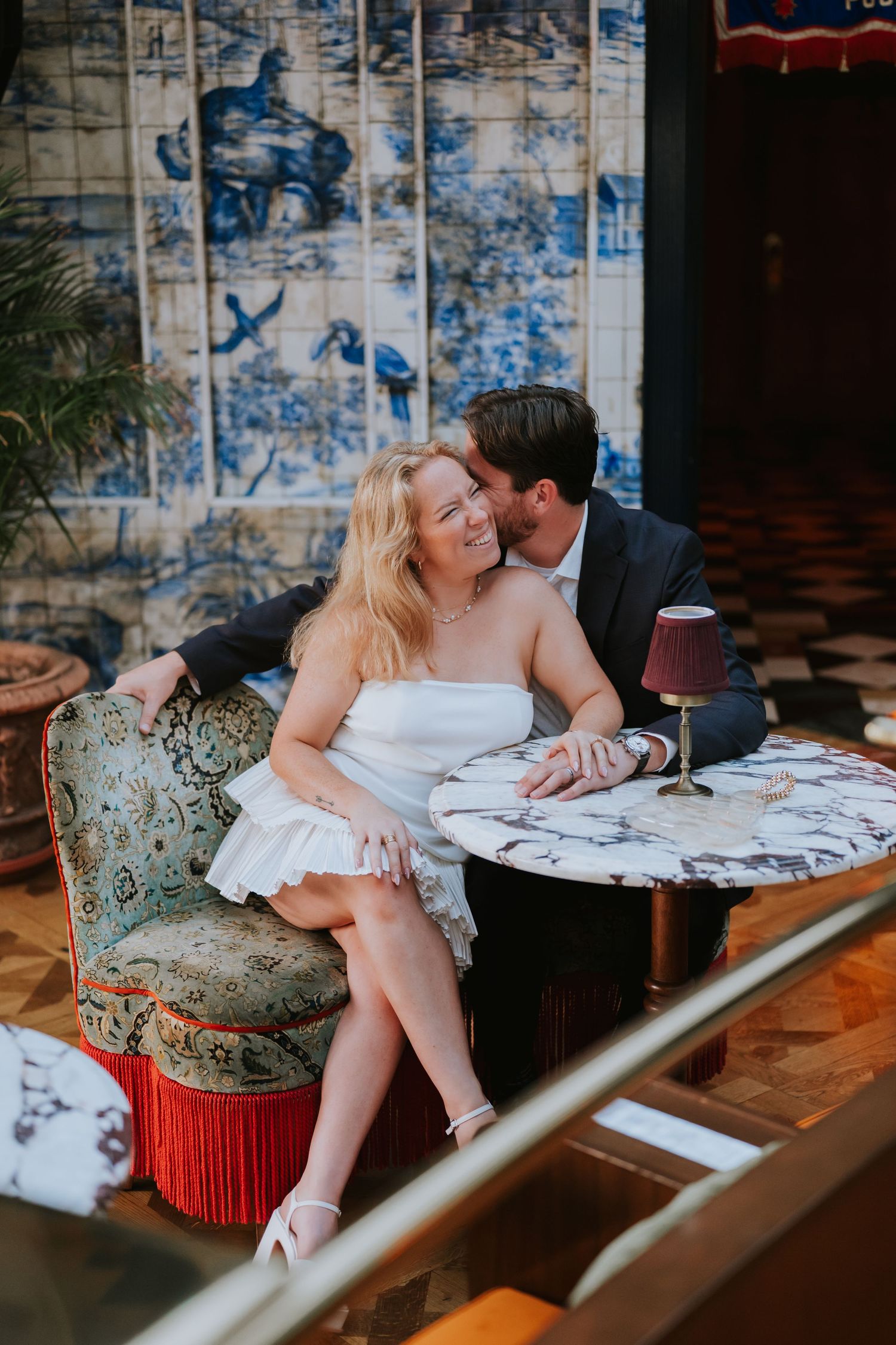 Groom kisses bride on the cheek as she smiles while seated on an ornate vintage sofa.