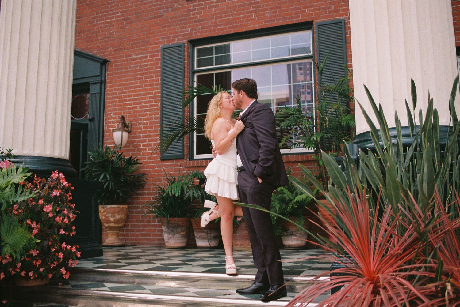 Couple sharing a romantic kiss outside a brick building surrounded by lush tropical plants.