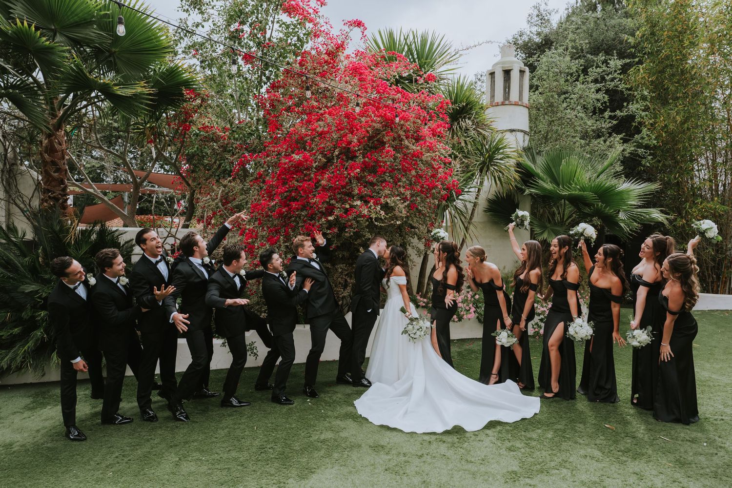Wedding party poses outdoors with bride and groom, groomsmen in black tuxedos and bridesmaids in black gowns near red bougainvillea.