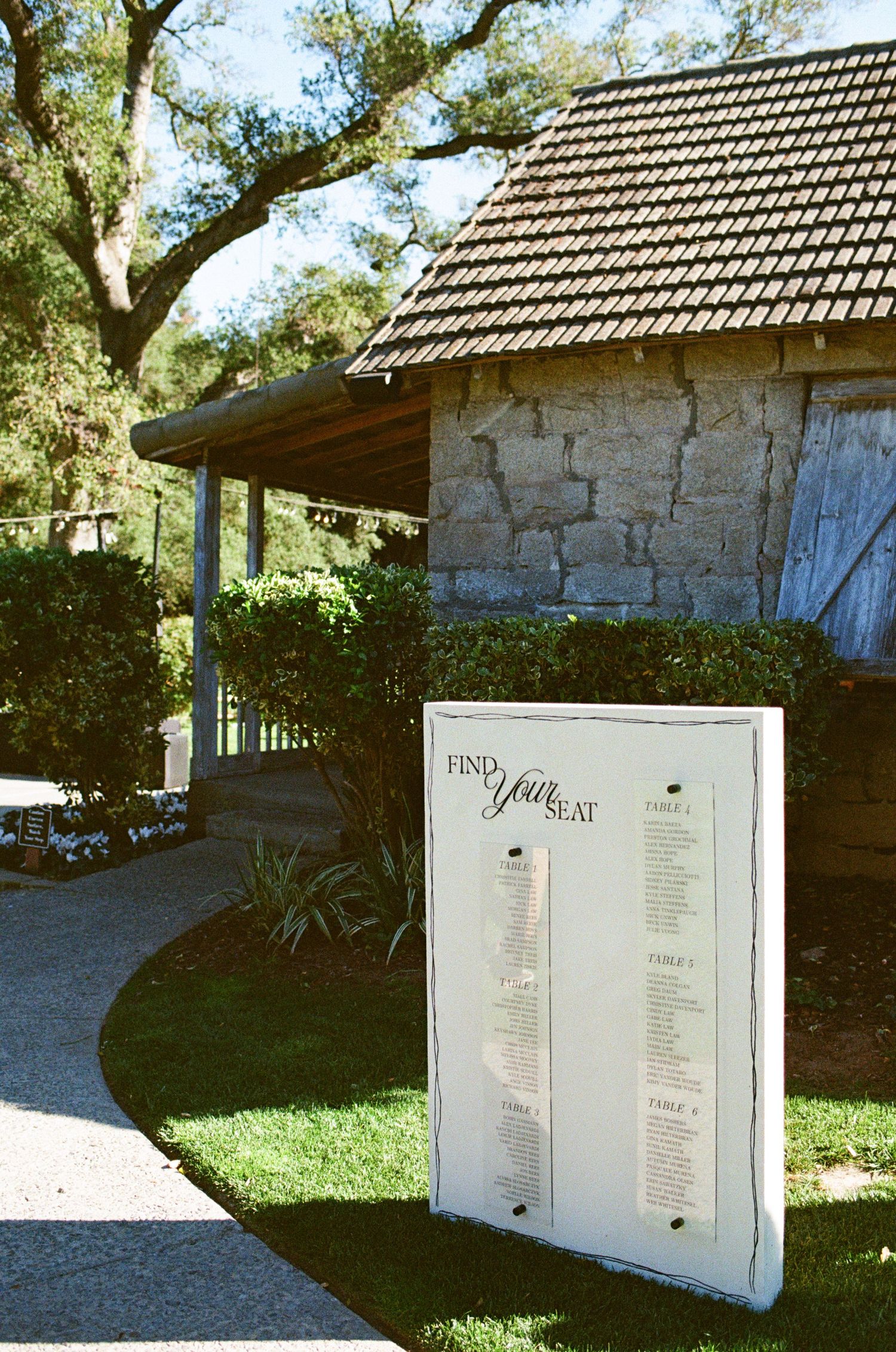 Outdoor wedding seating chart sign reading Find Your Seat displayed on white board near stone building and manicured garden path.