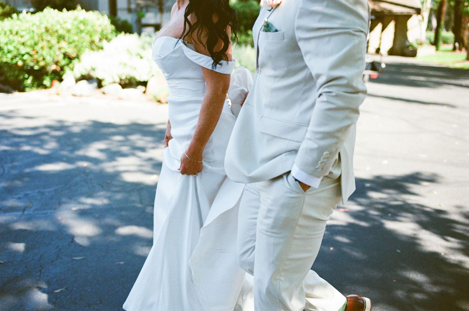 Bride in white off-shoulder gown and groom in beige suit walking hand in hand on a sunny path.