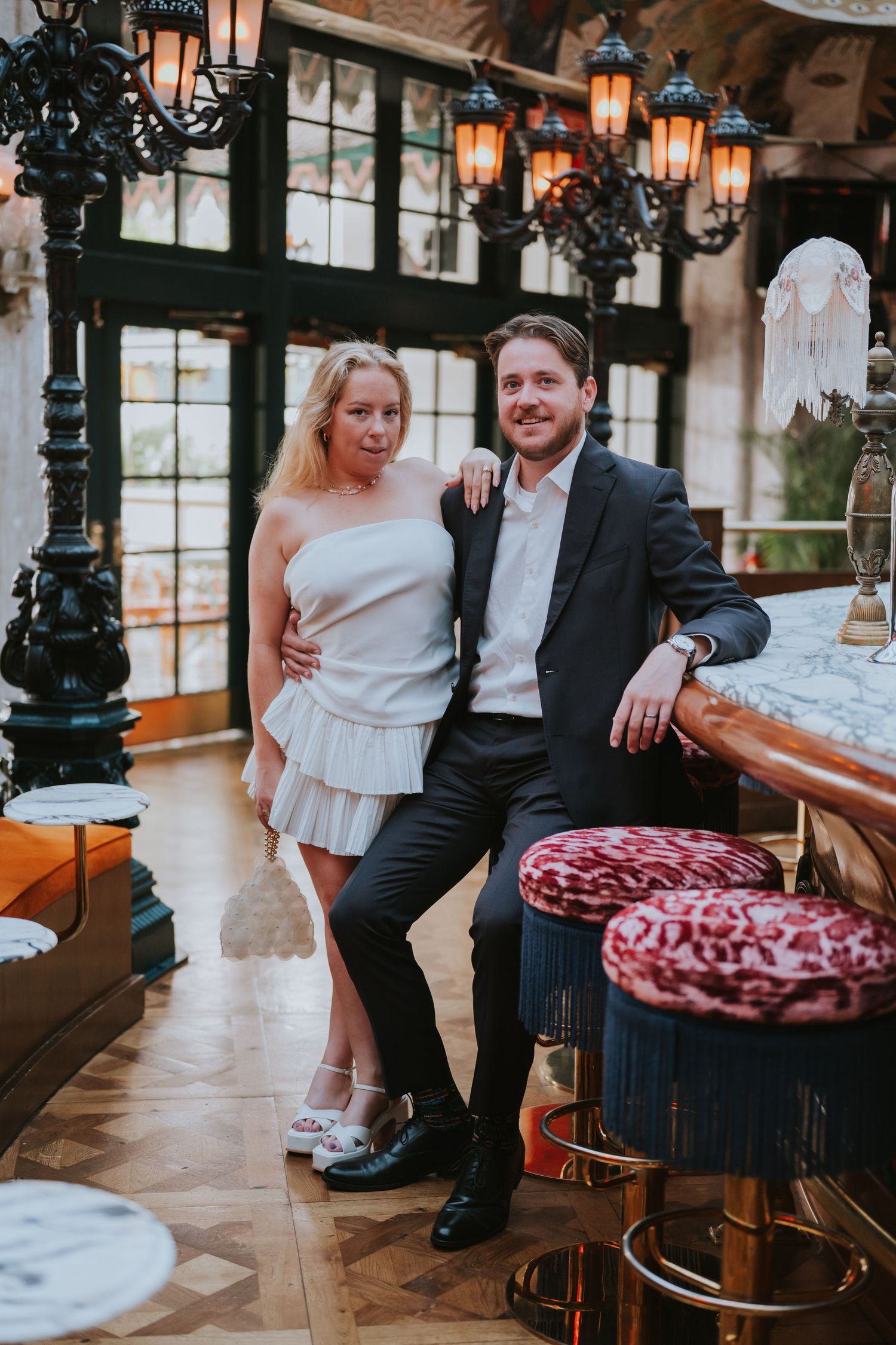 Couple posing in elegant bar, woman in white dress, man in dark suit, ornate lanterns and velvet stools.