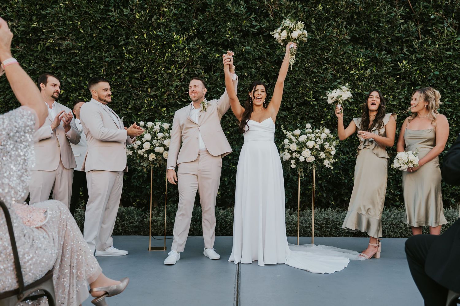 Wedding party celebrates joyfully on outdoor patio with white floral wall backdrop and elegant decor.