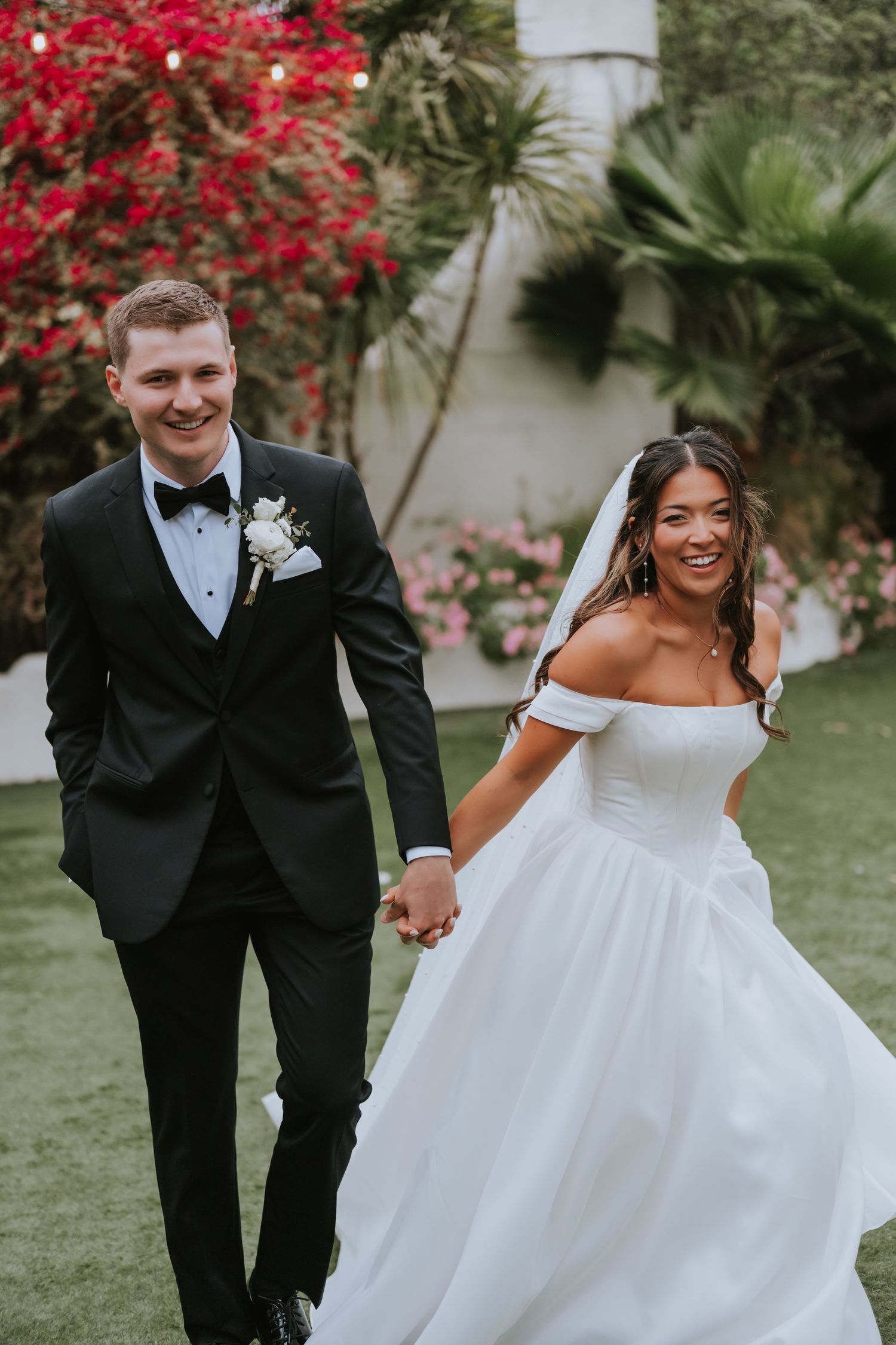 Smiling bride in off-shoulder white gown and groom in black tuxedo hold hands walking outdoors with floral backdrop.