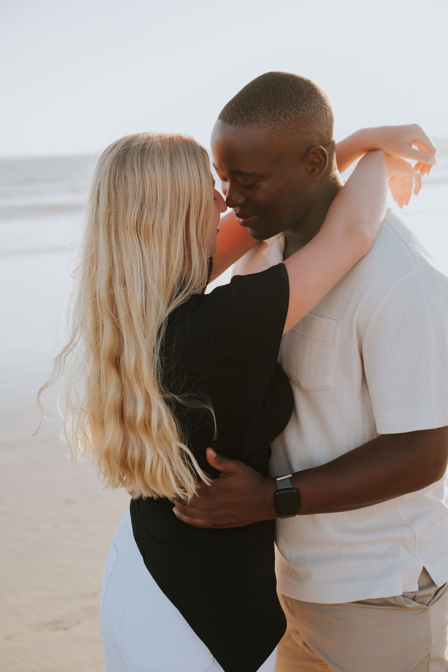 Interracial couple embracing romantically on a beach, woman in black dress with long blonde hair, man in cream shirt.
