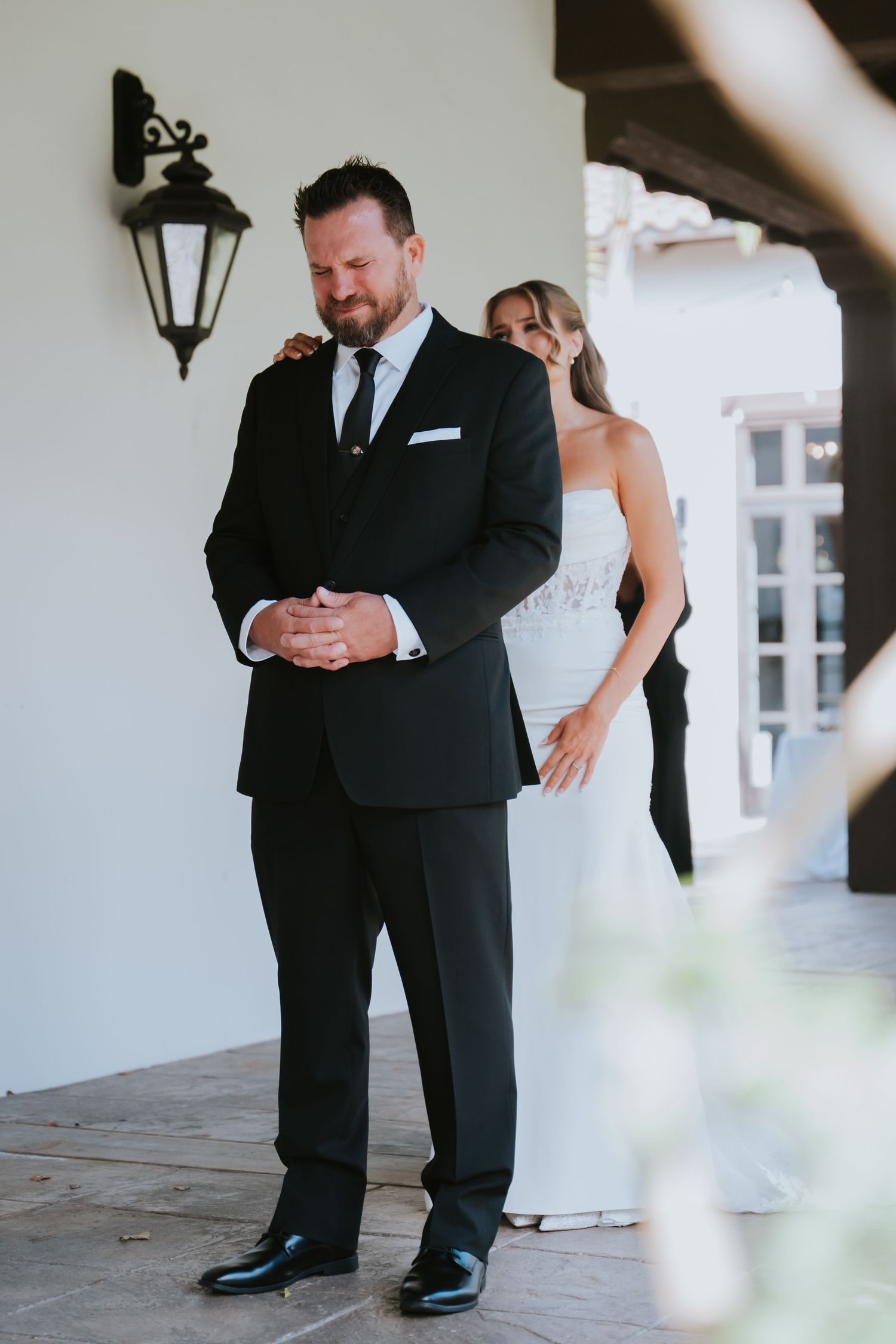 Groom in black tuxedo experiencing emotional first look moment with bride in white lace wedding dress on outdoor patio.