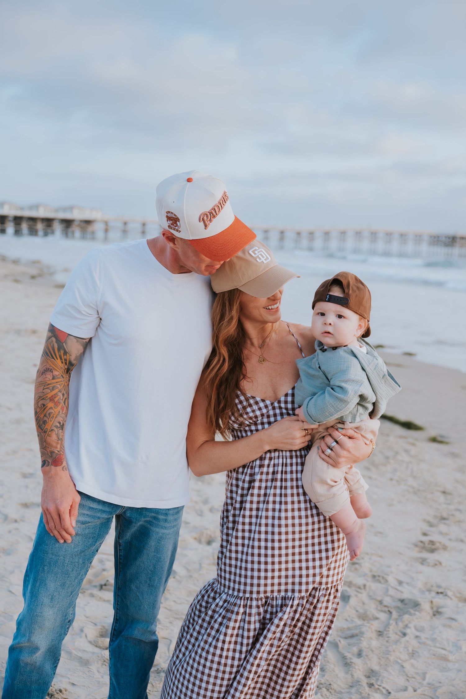 A couple with tattoos and baseball caps share a tender moment holding their baby on a sunny beach near a pier.
