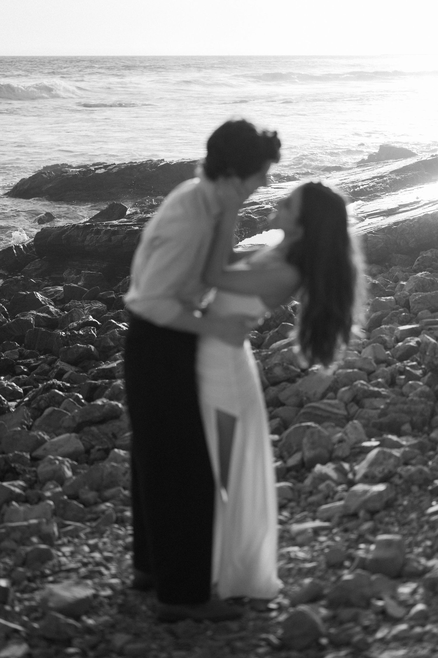 Black and white photo of couple embracing on rocky beach by ocean.