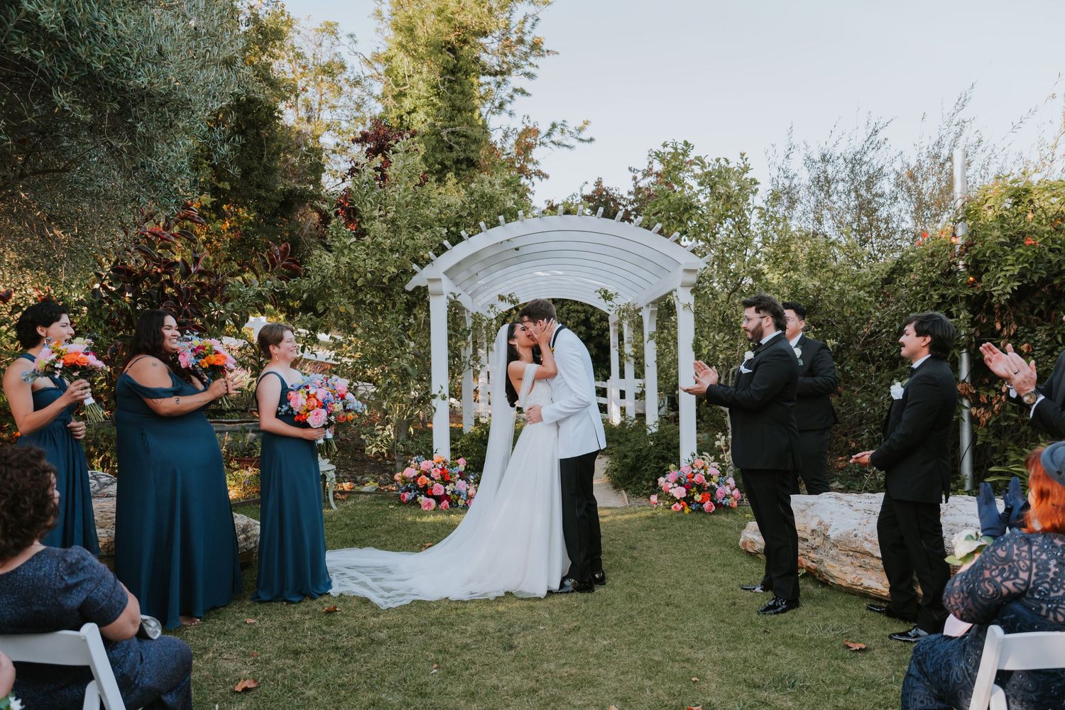 Outdoor garden wedding ceremony under white gazebo with wedding party in teal and black attire.