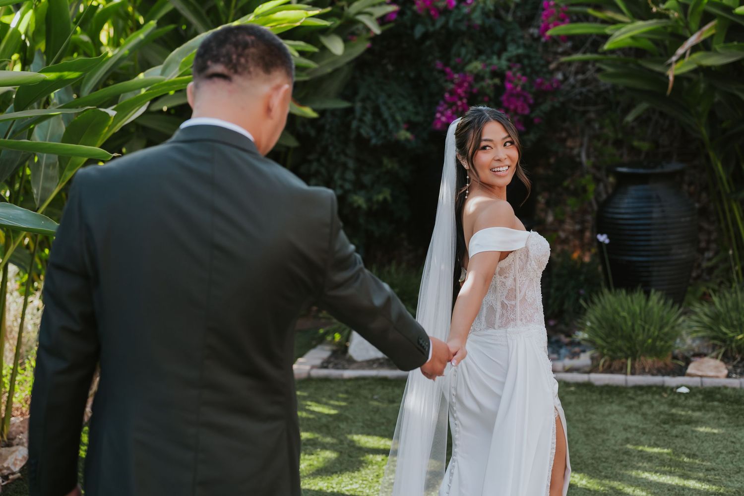A bride in a white dress poses with a veil in a garden setting during a wedding photoshoot.
