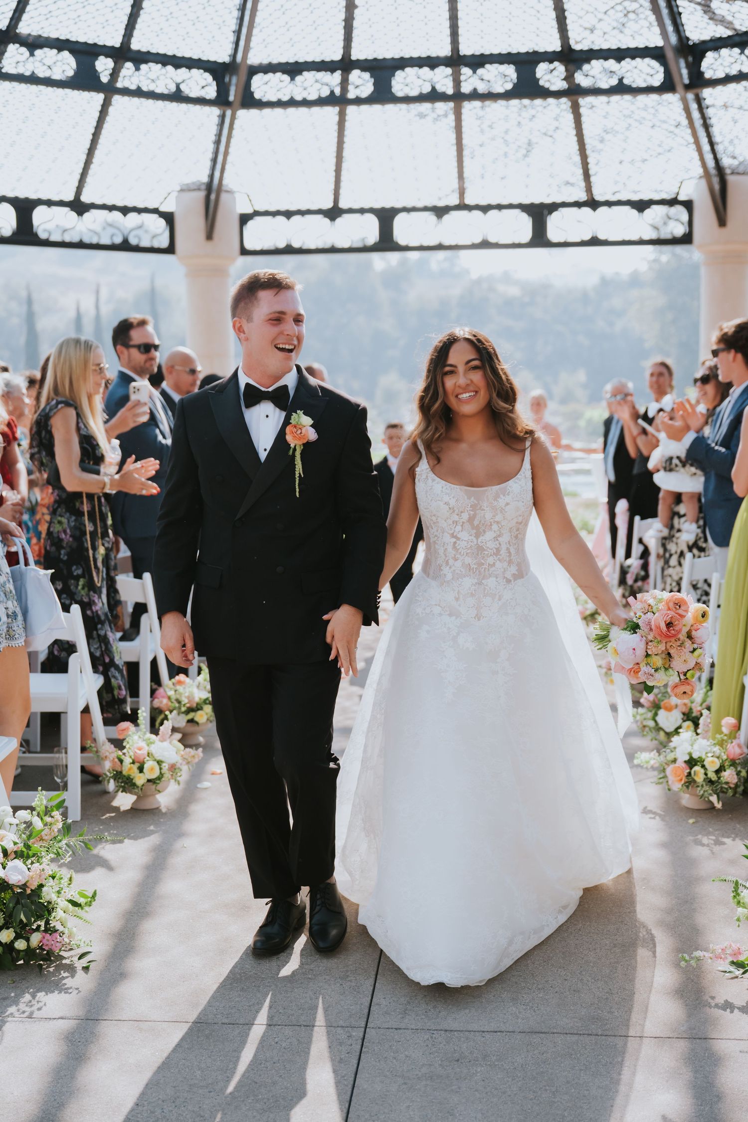 Bride and groom walk down the aisle hand in hand under a glass dome at an elegant outdoor wedding ceremony.