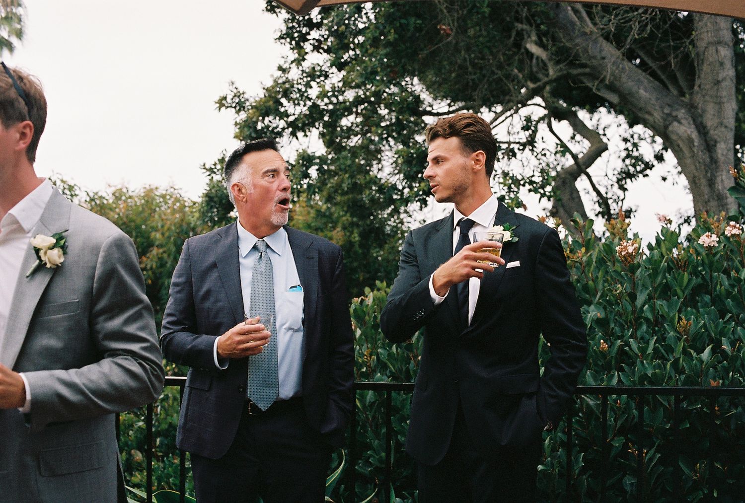 Two men in suits conversing at an outdoor wedding reception with greenery in the background.